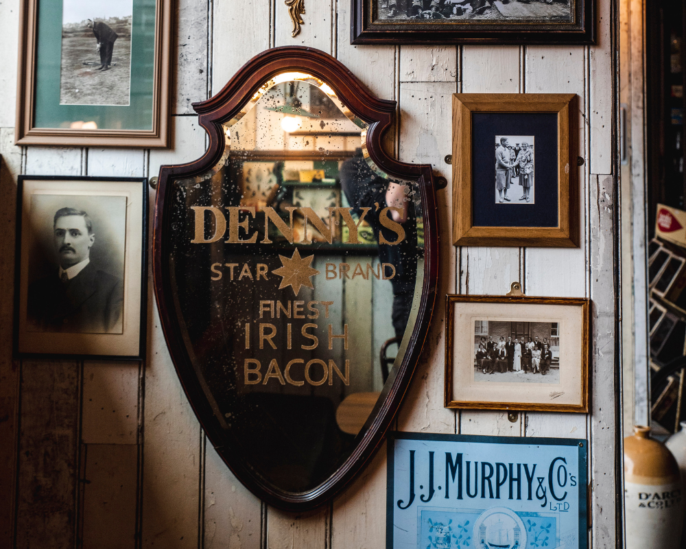 A wooden wall decorated with vintage framed photos and an ornate mirror with the text 'DENNY'S STAR BRAND FINEST IRISH BACON' written on it.