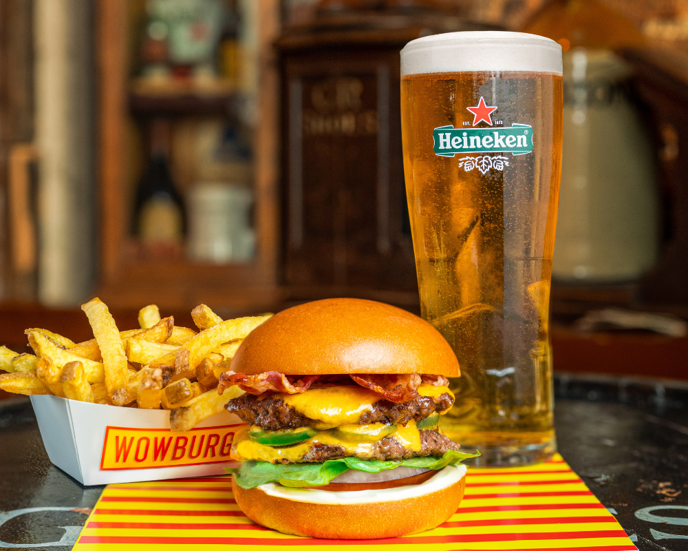 Burger with bacon, cheese, lettuce, tomato, and pickles on a bun, served with French fries on a tray labeled "WOWBURG", a tall glass of beer, and a pub interior in the background.