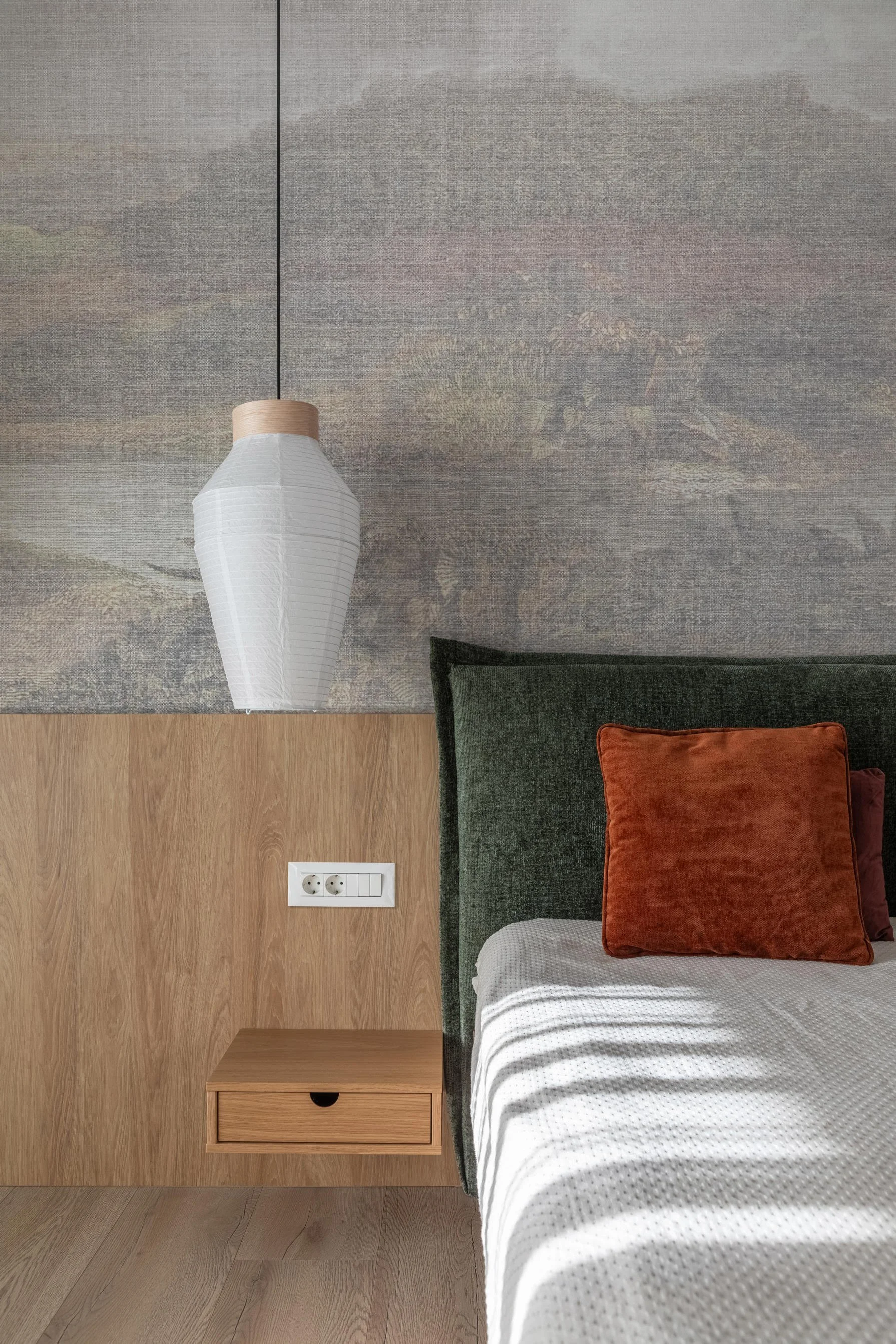 Bedroom with a bed covered in a white and gray dotted duvet, a green and black rug, an orange cushion, a wooden nightstand, a white paper lantern pendant light, and a wall socket, with a woven rug on the floor.