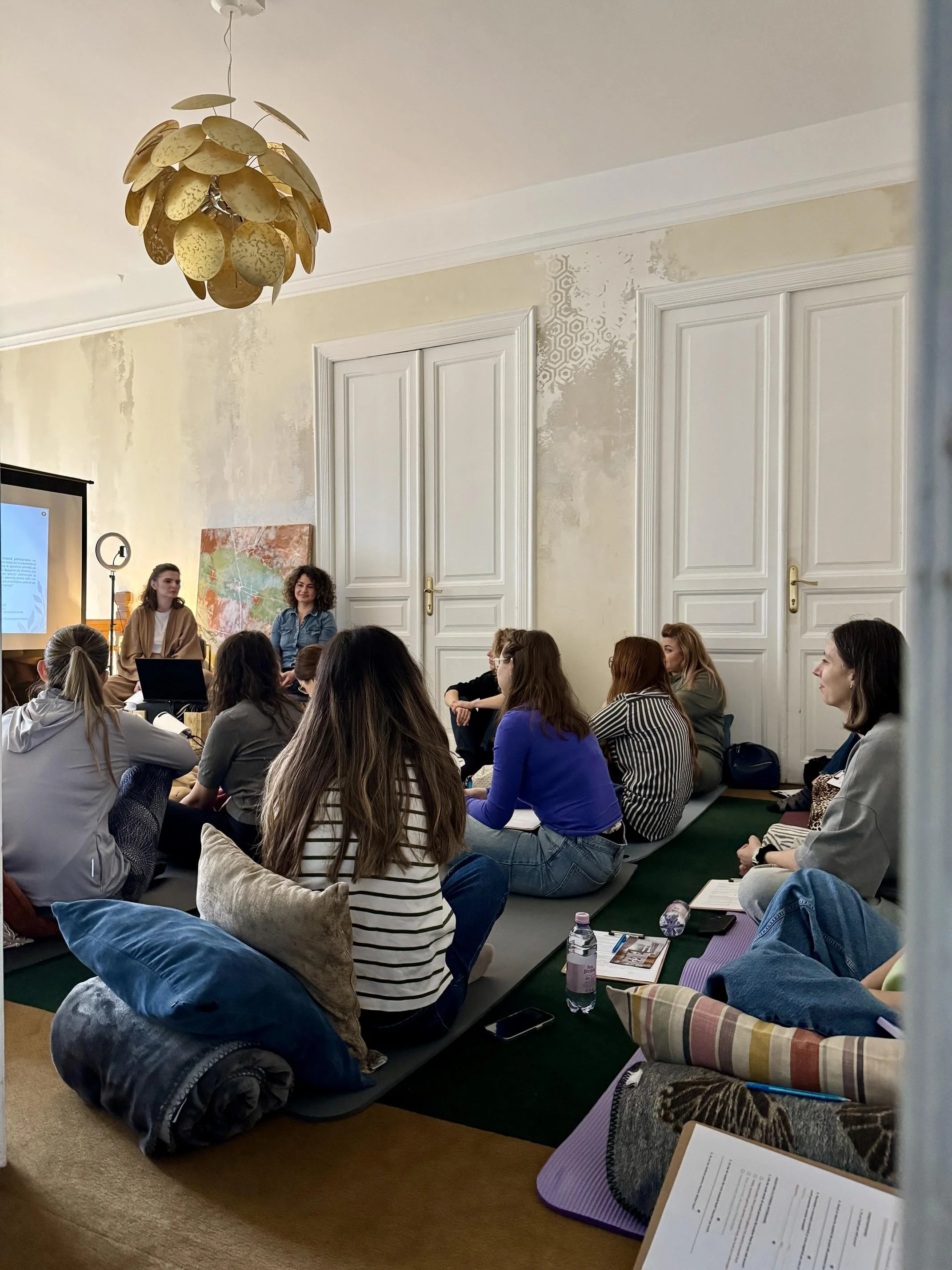 A group of people sitting on cushions and mats on the floor in a room, attending a presentation or workshop. Two women are sitting at the front near a presentation screen, one with curly hair smiling. The room has white walls, ornate doors, and a decorative chandelier hanging from the ceiling.