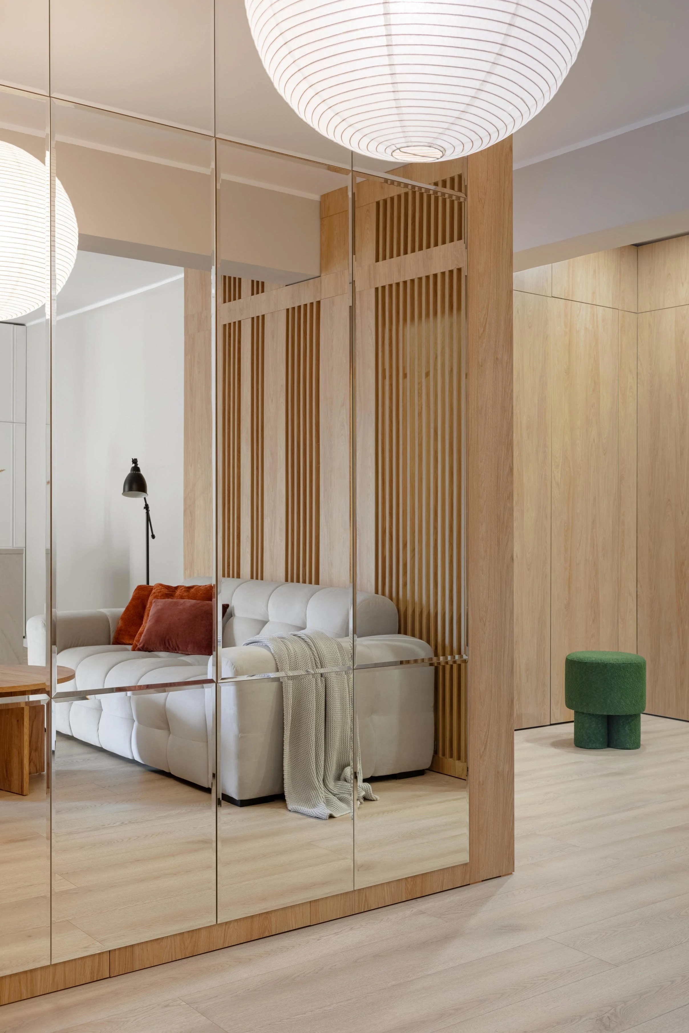 A living room with a white sofa with brown and pink pillows, a black floor lamp, a wooden side table, a large mirror reflecting part of the room, a wooden partition with slats, a white ceiling with a round paper lantern, and a green ottoman.