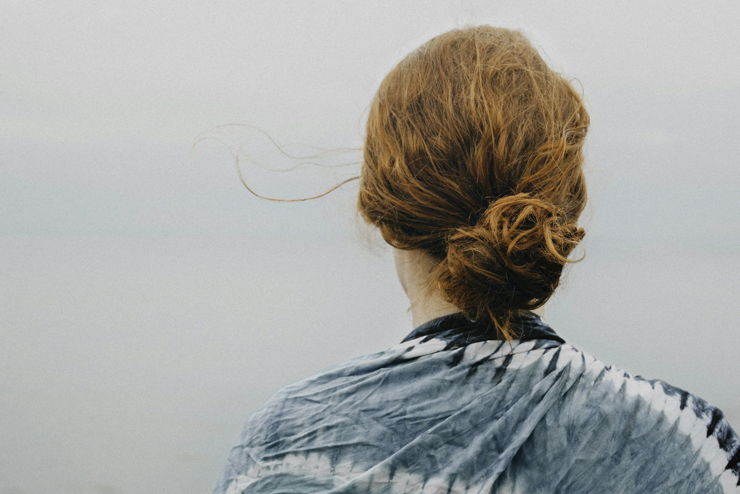 A person with red, curly hair seen from behind, wearing a blue and white tie-dye shirt, standing outdoors against a light sky.