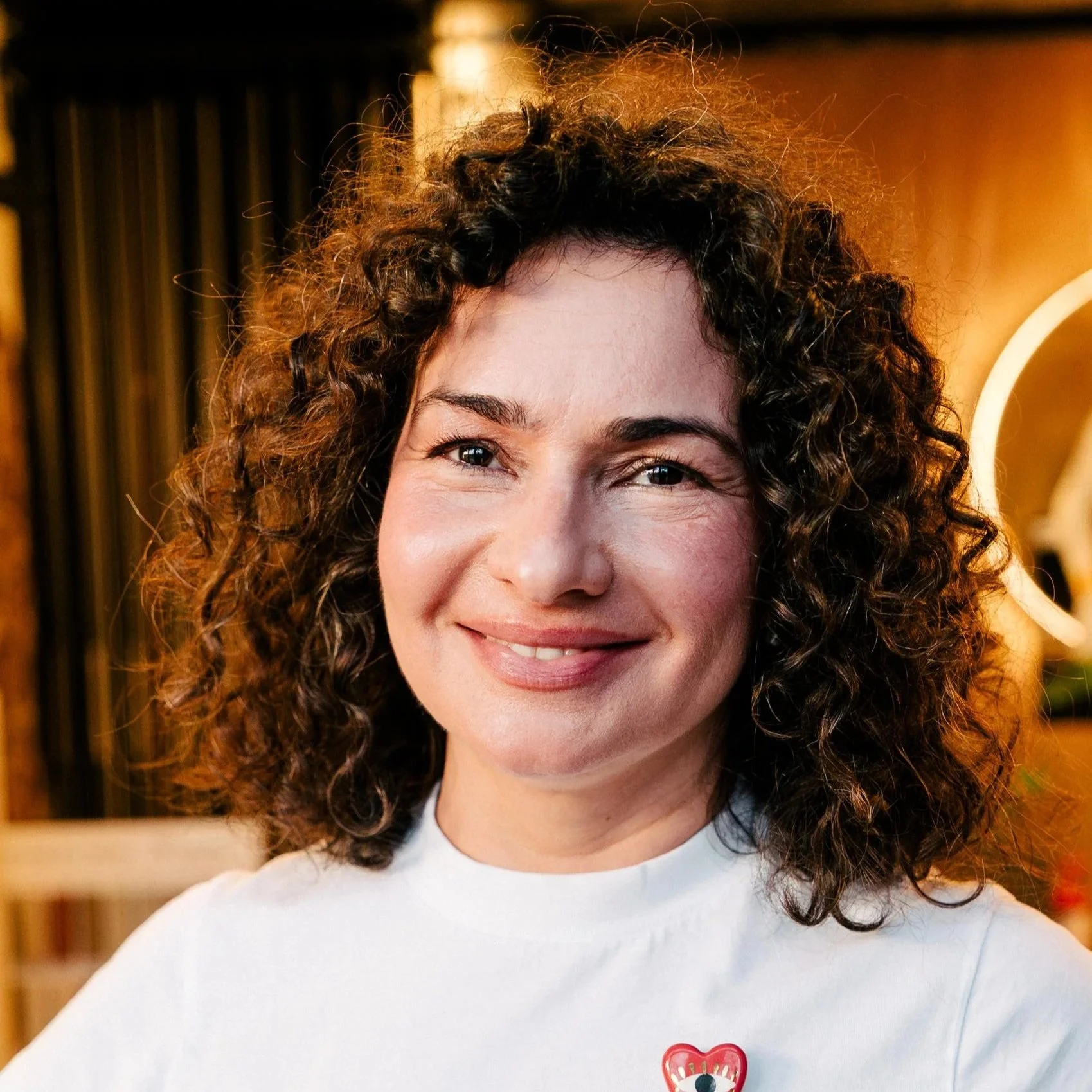 Close-up of a woman's smiling face with curly hair, wearing a white shirt with a red heart pin, in an indoor setting with a blurred background.