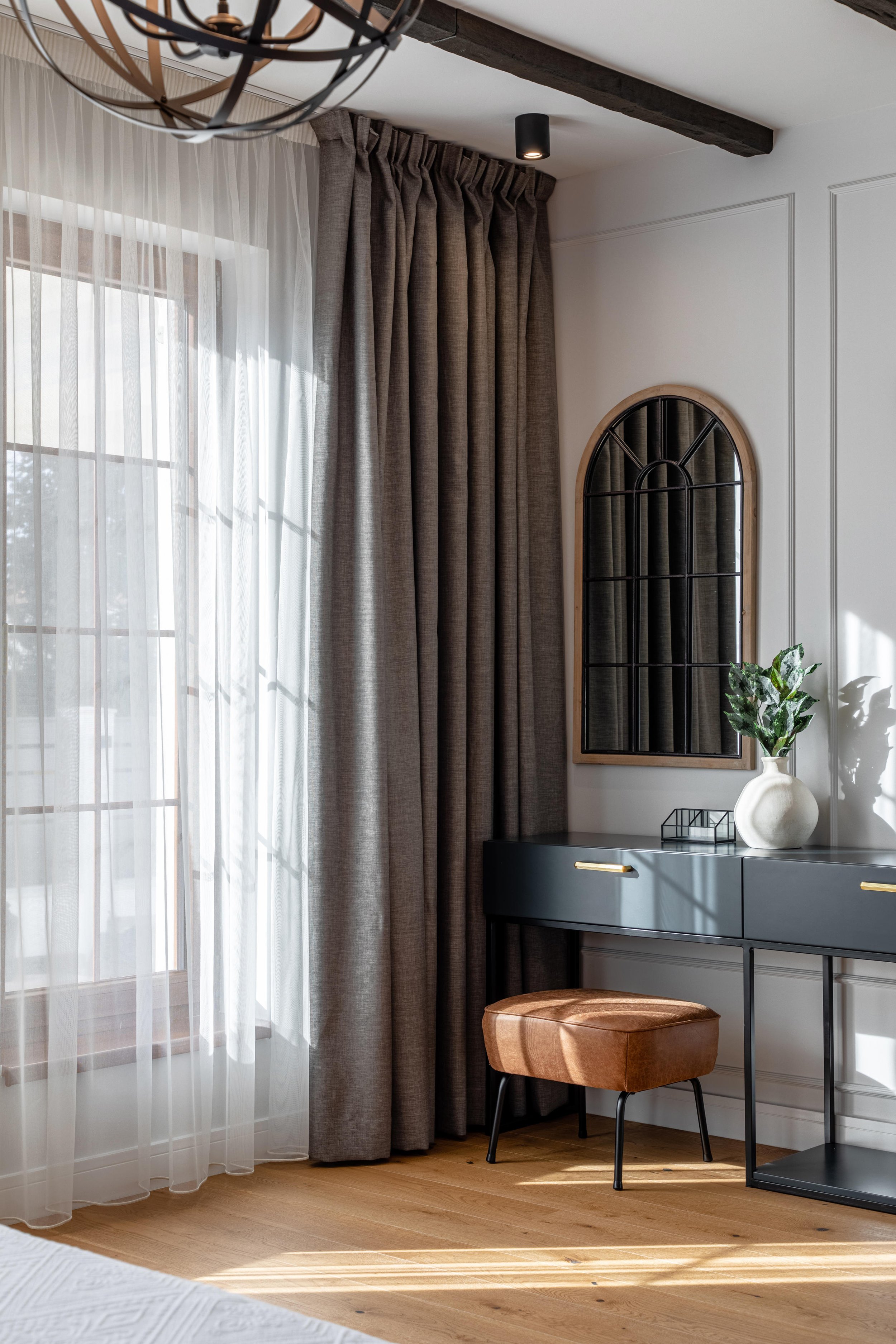 A corner of a modern room featuring a window with white sheer curtains and darker gray drapes, a dark grey console table with gold handles, a decorative mirror with a black grid pattern and wooden frame, a potted plant, a small black tray, and a tan ottoman with black legs. The room has wood flooring and white walls with decorative molding, and a dark ceiling beam with a ceiling light.