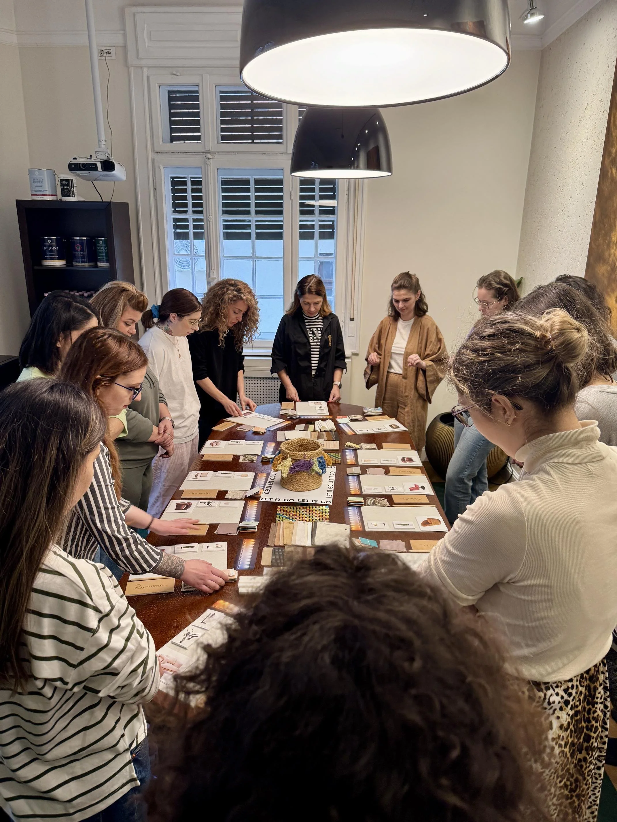 A group of people gathered around a table with various cards and papers, engaged in an activity or discussion in a well-lit room with large windows and hanging lights.