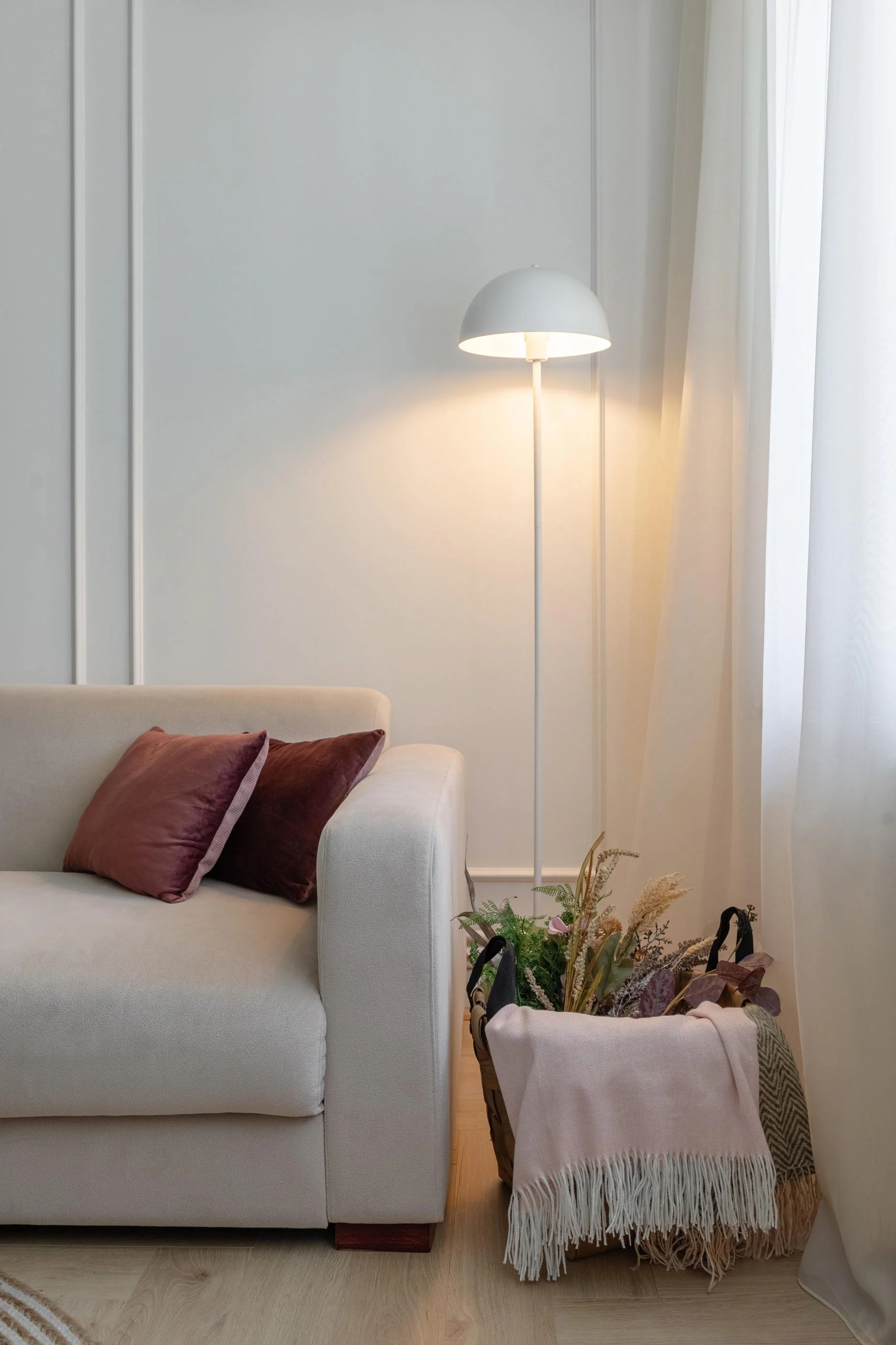 A cozy living room corner with a white upholstered sofa, two burgundy pillows, a tall white floor lamp, a basket with an assortment of dried flowers and plants, and white curtains covering the window.