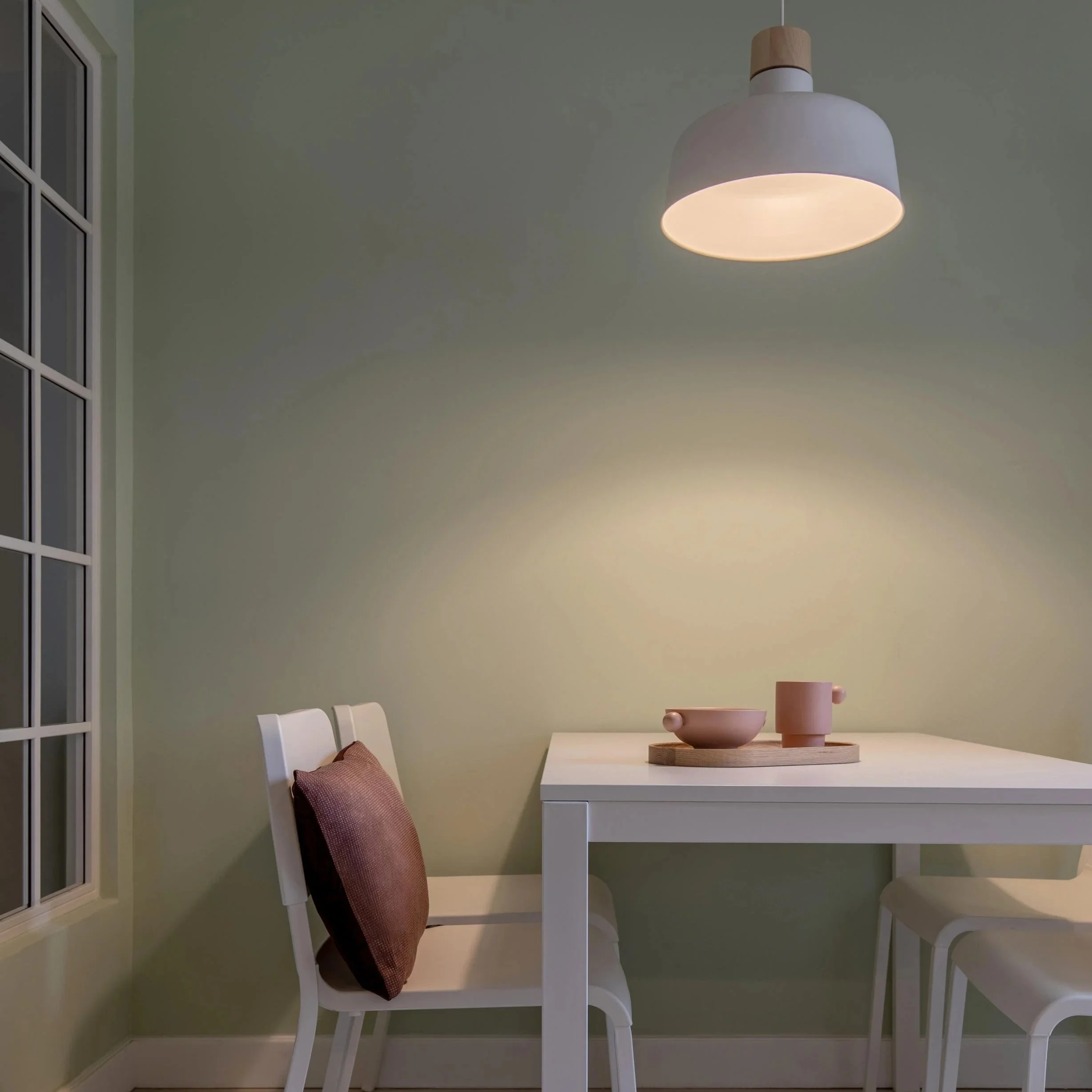 A white dining table with a pink vase and tray, two white chairs, and a cushioned chair with a pink pillow, in a dining corner with a green wall and a window.