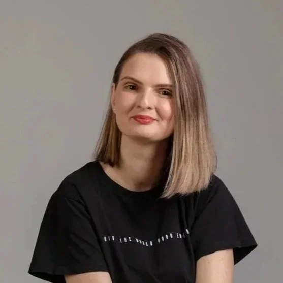 A young woman with shoulder-length blonde hair wearing a black t-shirt, smiling softly, against a neutral gray background.