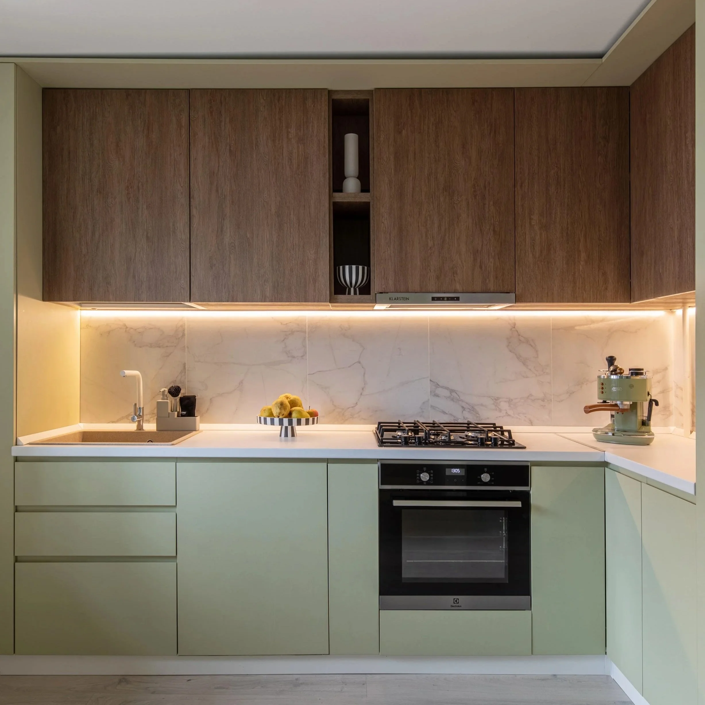 Modern kitchen with light green lower cabinets, wooden upper cabinets, marble backsplash, and built-in oven. Countertop has a bowl of lemons, a sink, and small appliances.