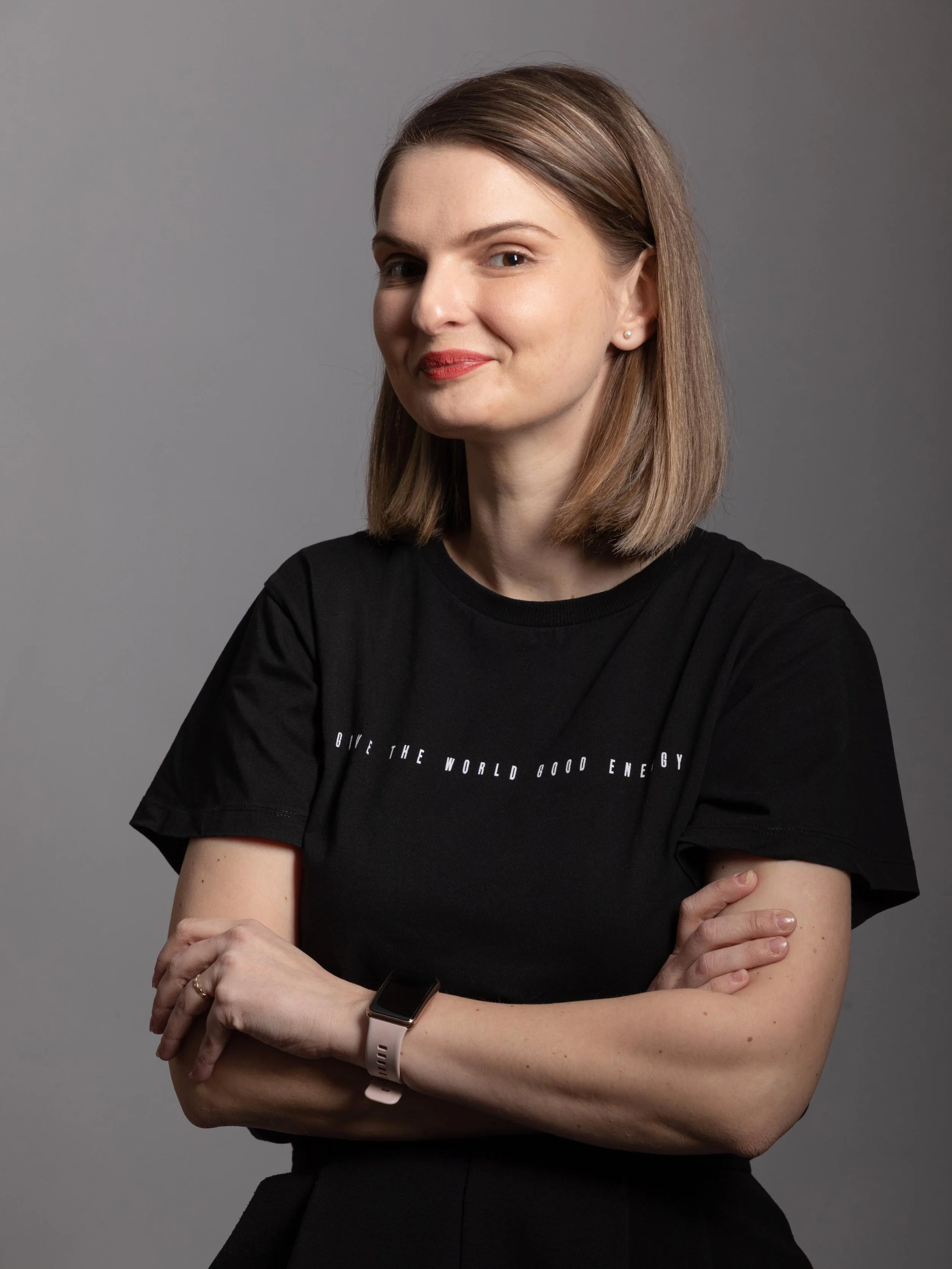 A woman with shoulder-length brown hair, wearing a black T-shirt with text, cross-armed, smiling gently, against a gray background.