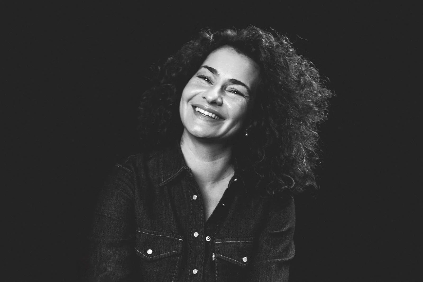 Black and white photo of a woman with curly hair, smiling and wearing a denim jacket.