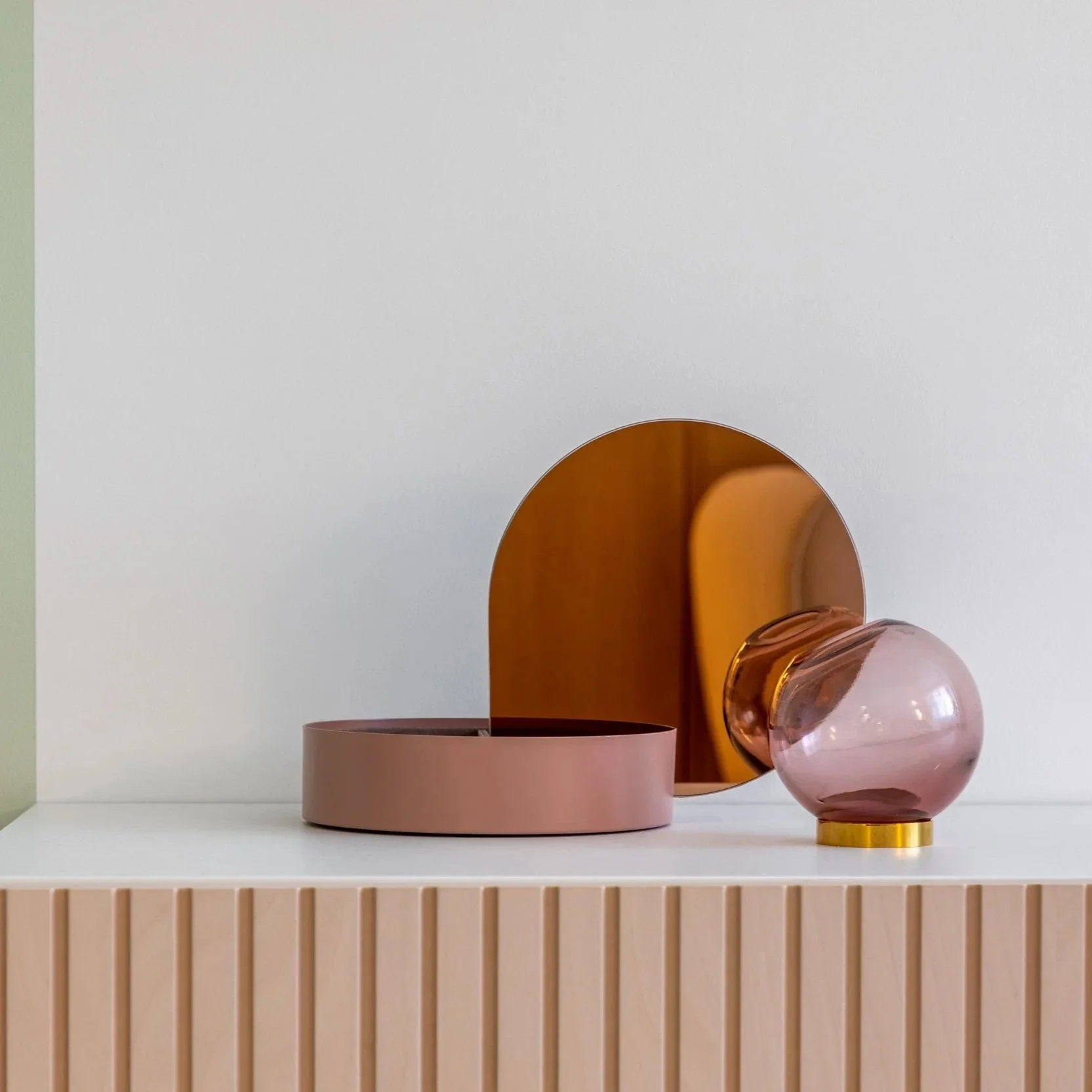 Decorative objects on a shelf including a pink bowl, a copper-colored oval mirror, a pink spherical object with a gold base, and a light-colored vertical slatted detail on the front of the shelf.