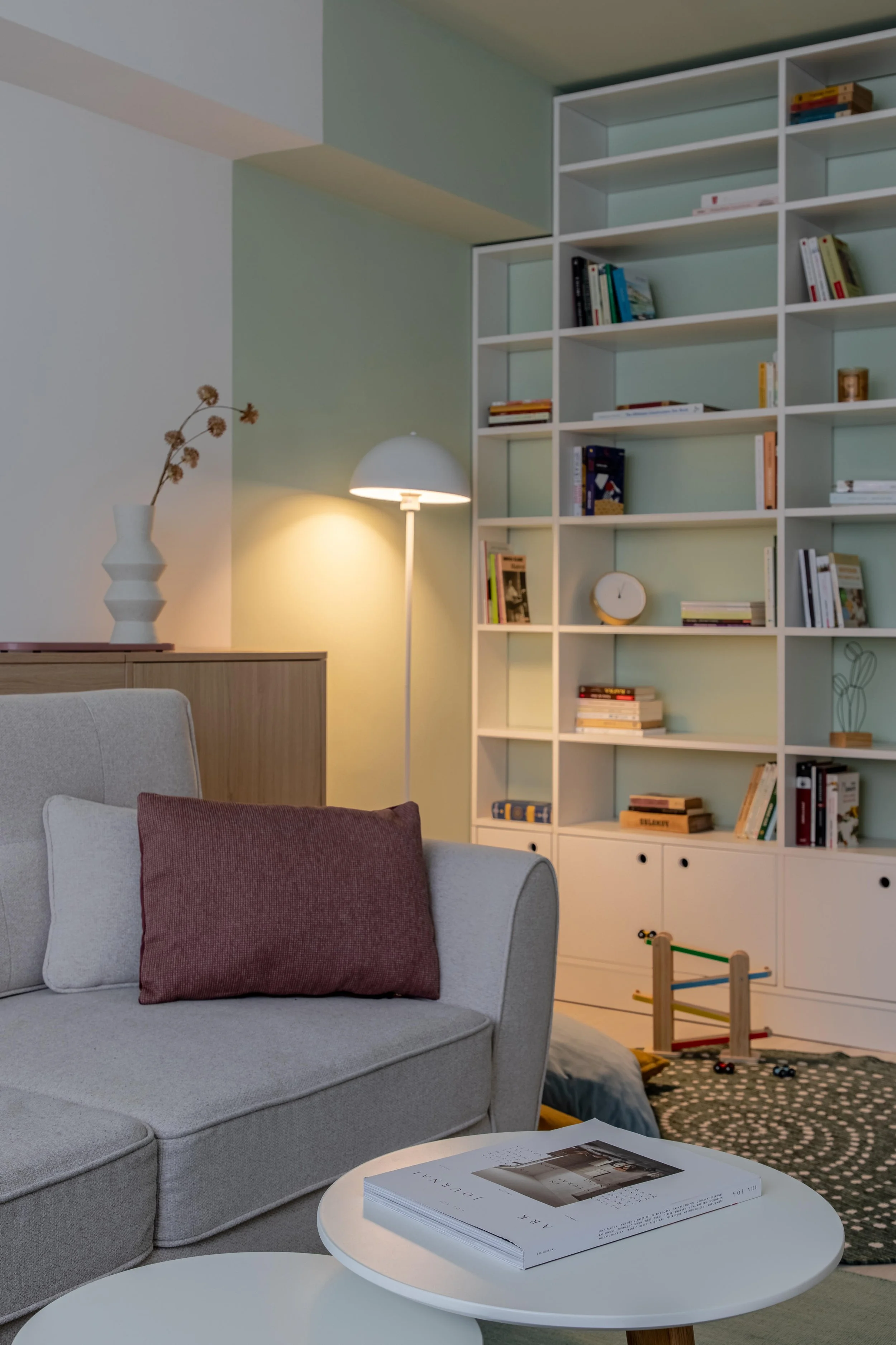Living room with a beige sofa, a red pillow, white coffee table with a stack of magazines, a white bookshelf filled with books, a floor lamp, a decorative vase with dried flowers, and a children's wooden toy in a patterned rug.