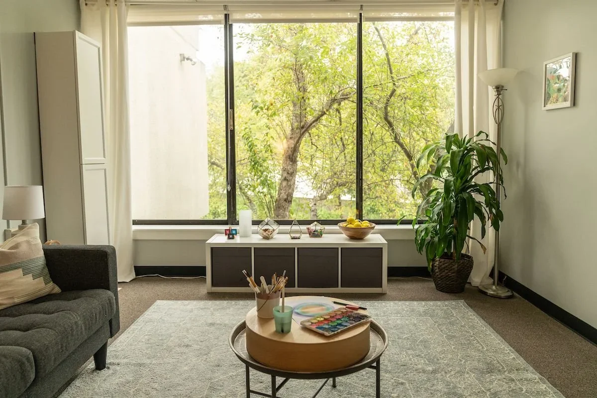 Modern therapy room with natural light, sofa, side table, plants, and calming artwork in a psychology clinic setting.