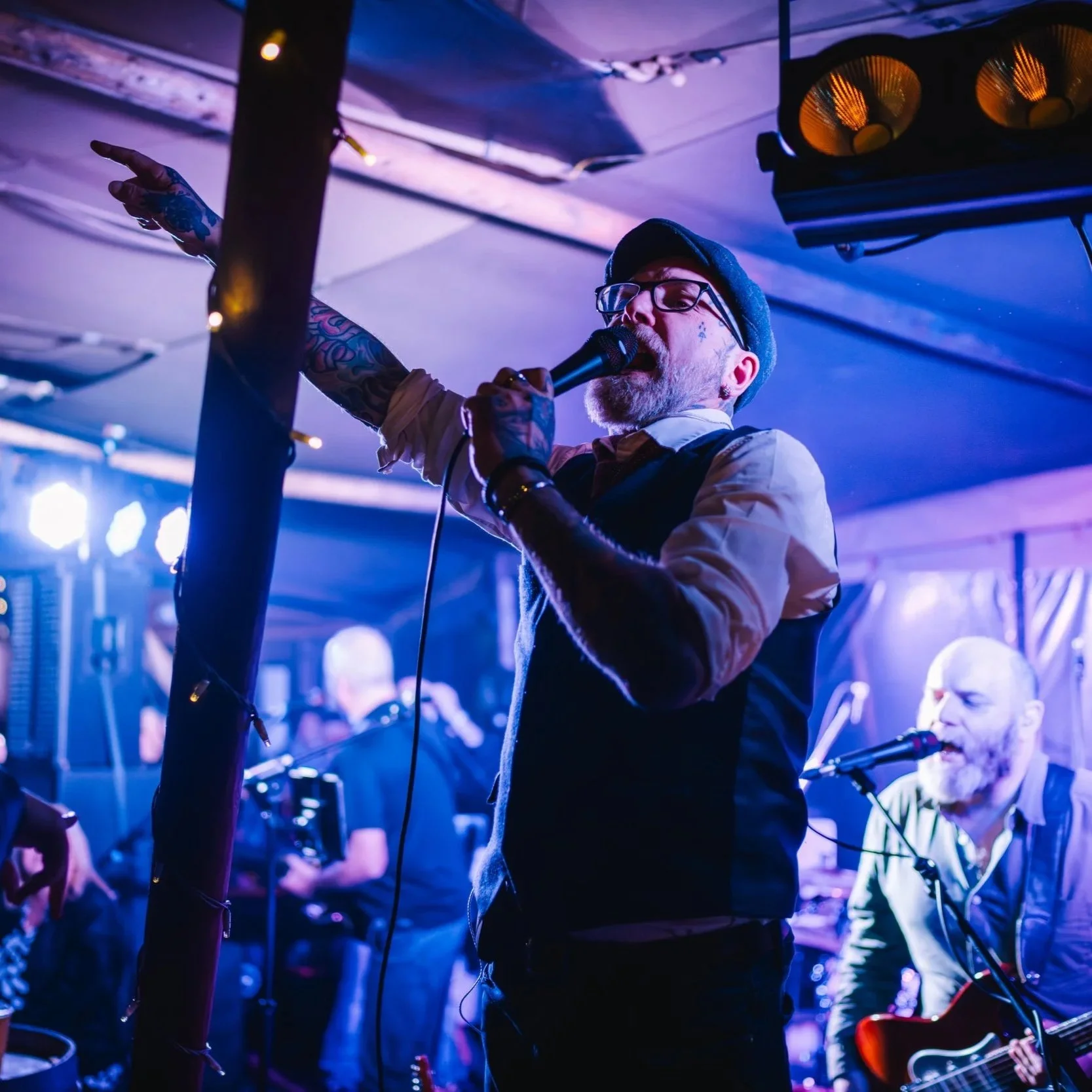 A man with glasses, tattoos, and a beret singing into a microphone on stage with purple and blue lighting, other musicians and instruments behind him.