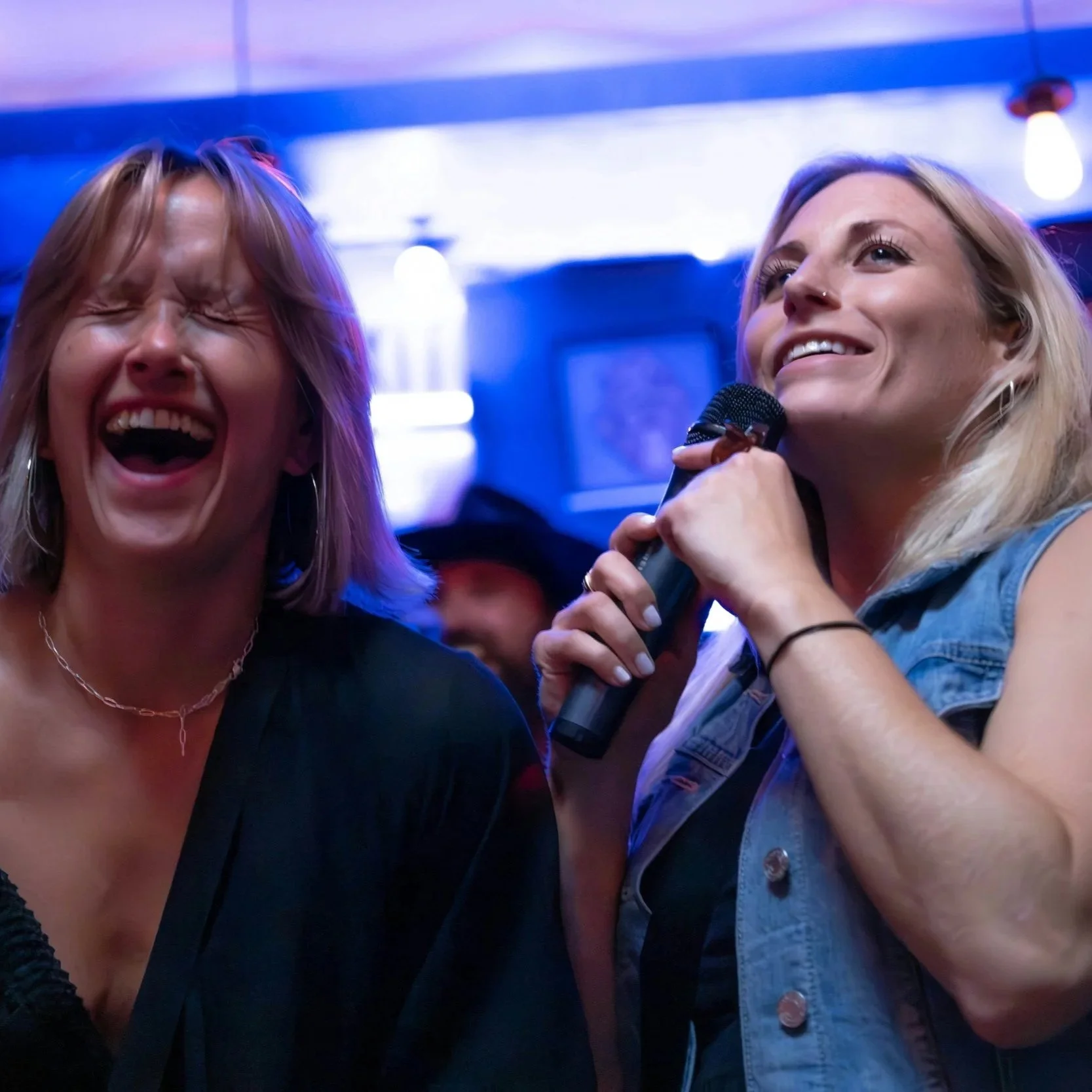 Two women at a social event, one laughing with eyes closed and mouth open, and the other speaking into a microphone, both with light hair, in a dimly lit room with blue and purple lighting.