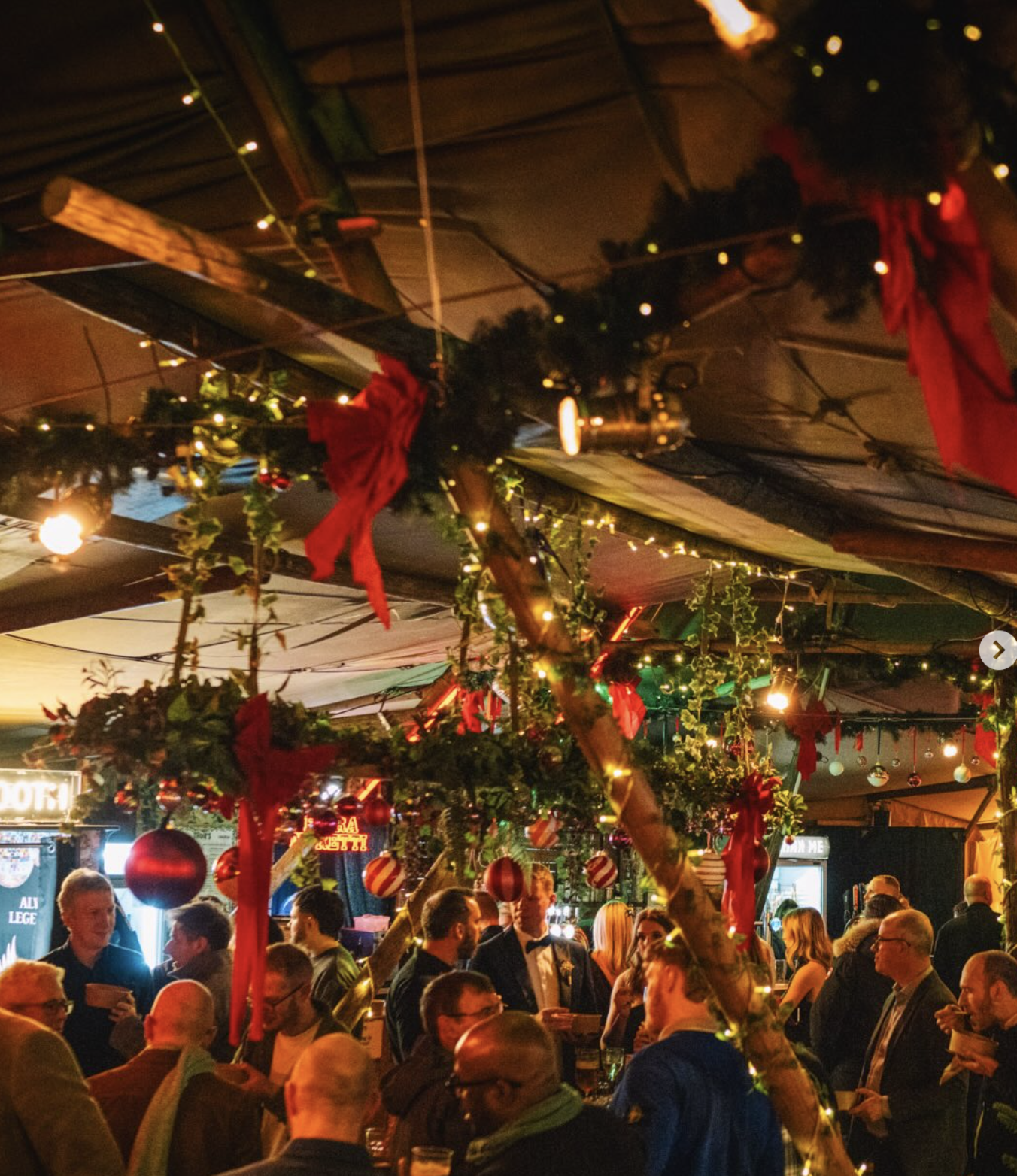 A crowded indoor party or celebration with people socializing, decorated with Christmas ornaments, red ribbons, and string lights hanging from the ceiling, creating a festive atmosphere.