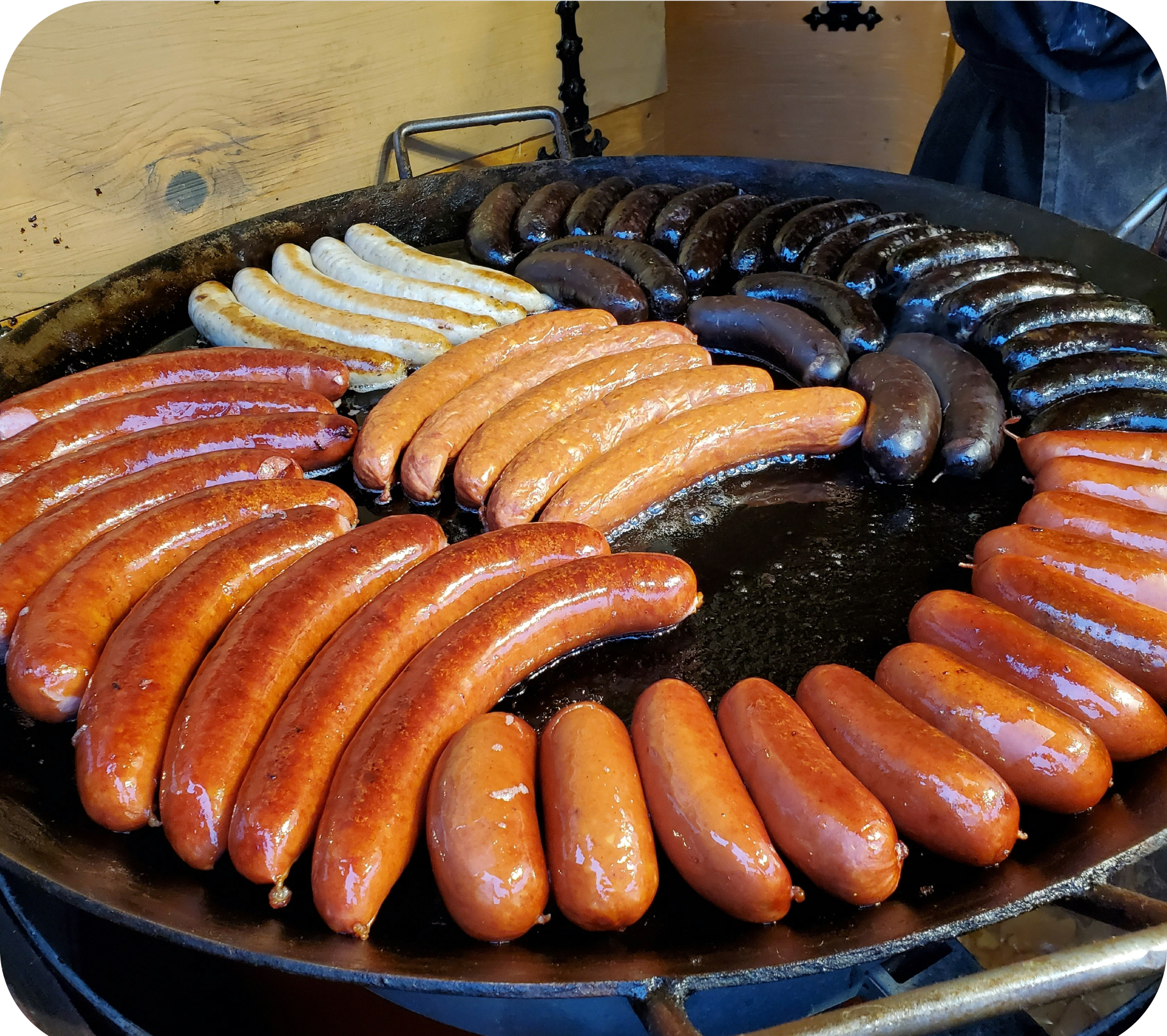 Assorted sausages being grilled on a cast iron skillet, with some sausages appearing more cooked and browned while others are lighter in color.