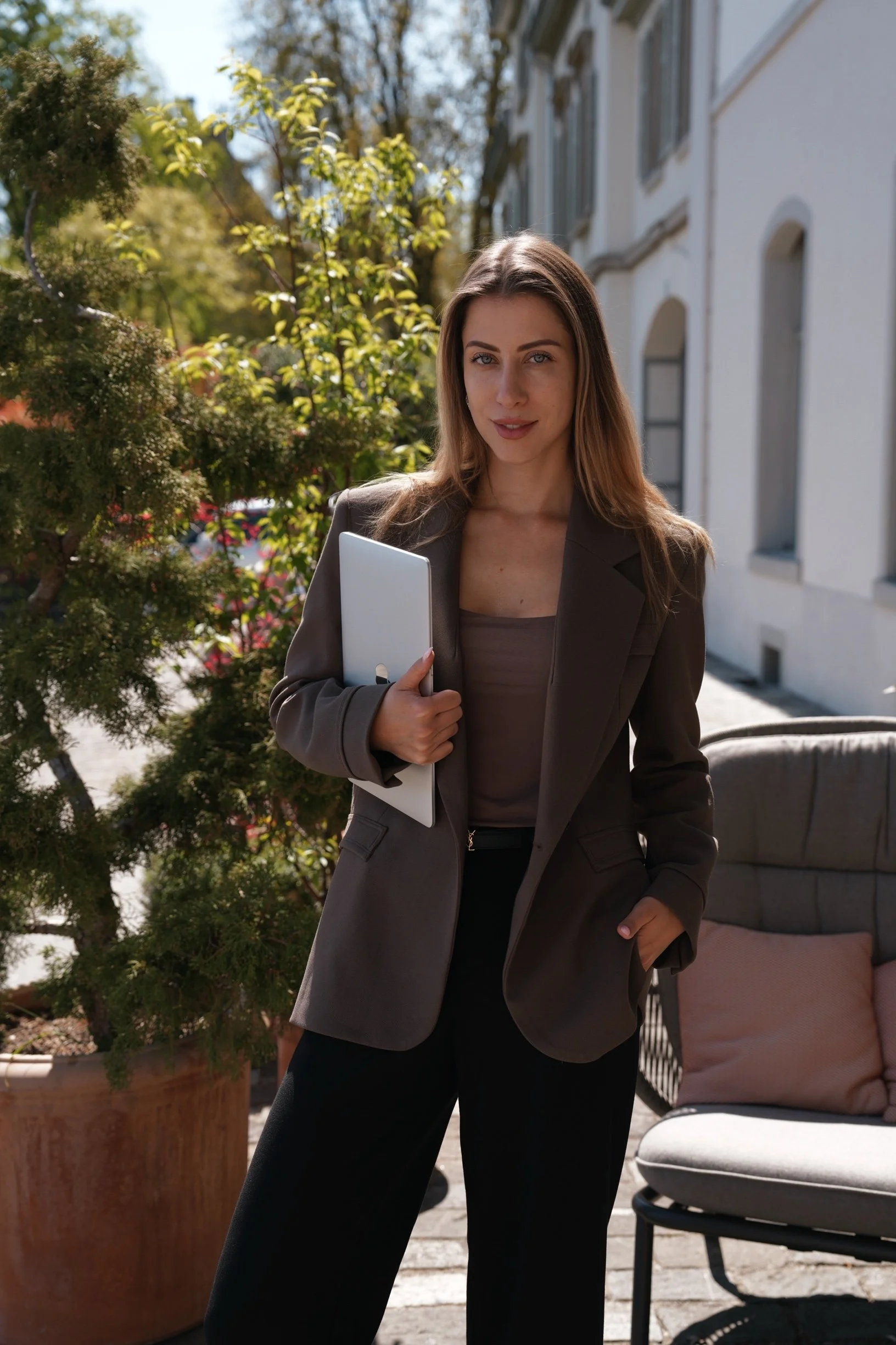 Founder of Secluded Agency. Young sophisticated woman wearing a business outfit - blazer and black pants, standing outdoors on a sunny day holding a laptop and ready to work.