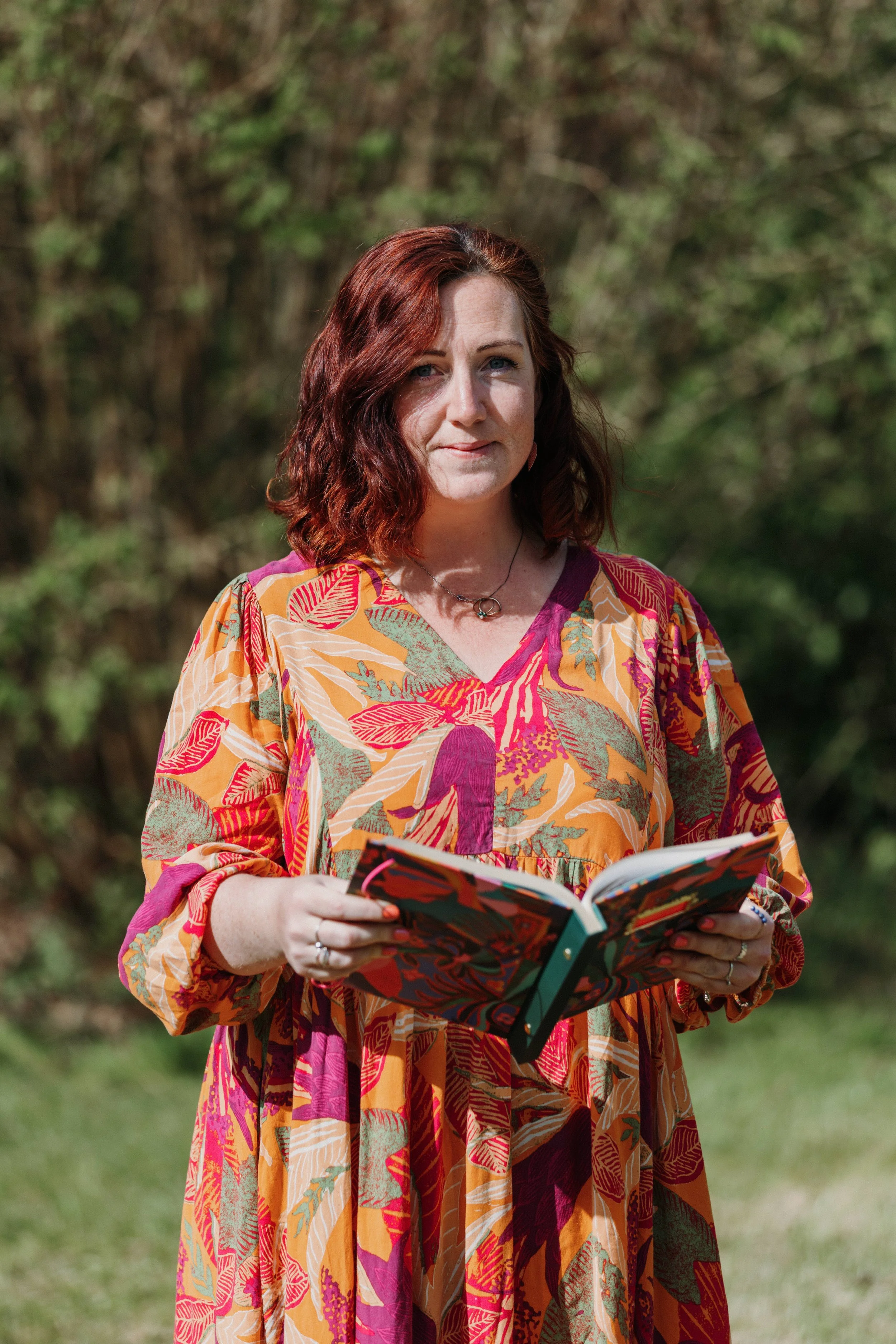A woman with red hair wearing a colorful floral dress is holding an open book outdoors with a background of trees and greenery.