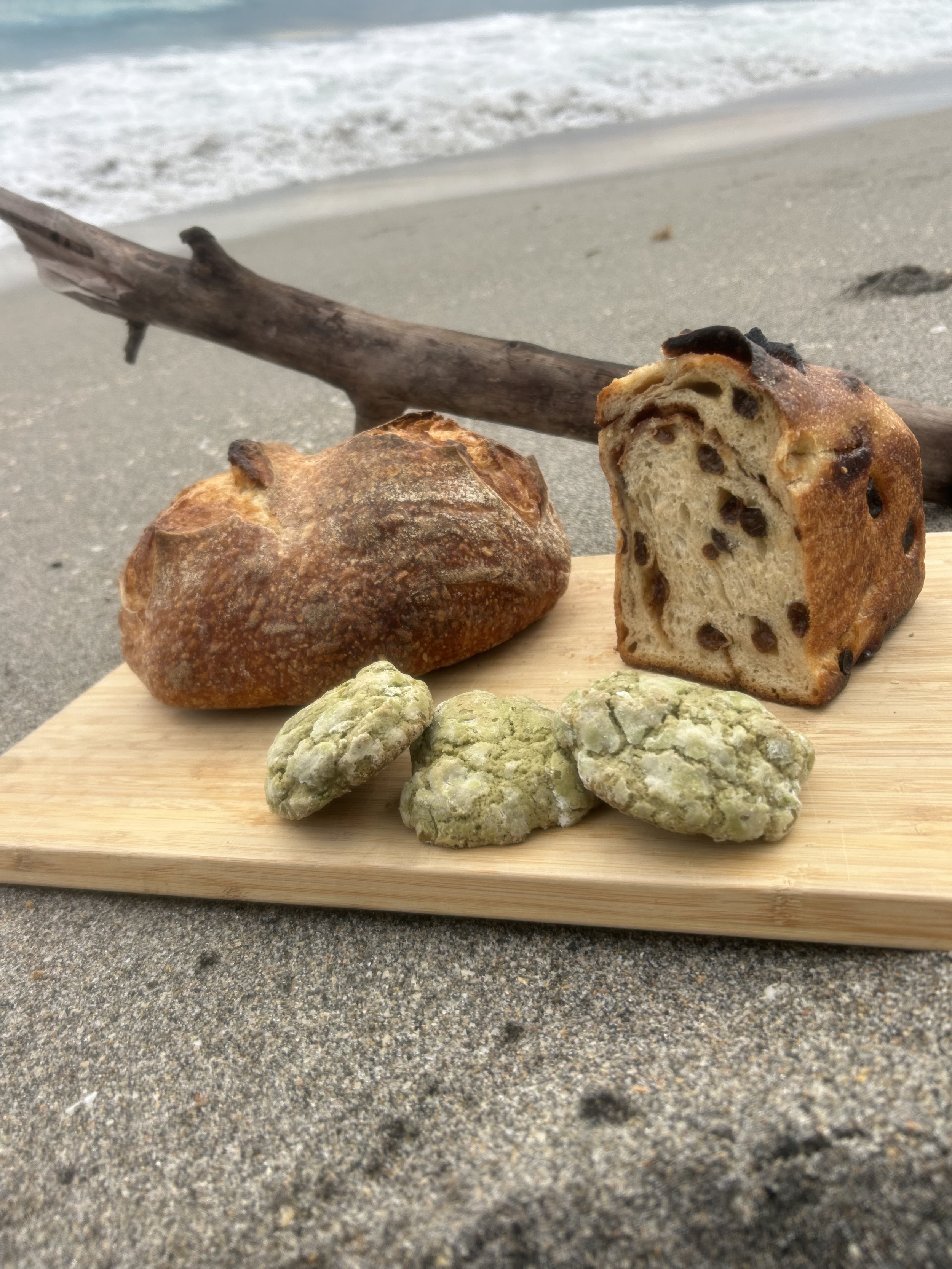 Bread and cookies placed on a wooden plank on a sandy beach, with driftwood and ocean waves in the background.