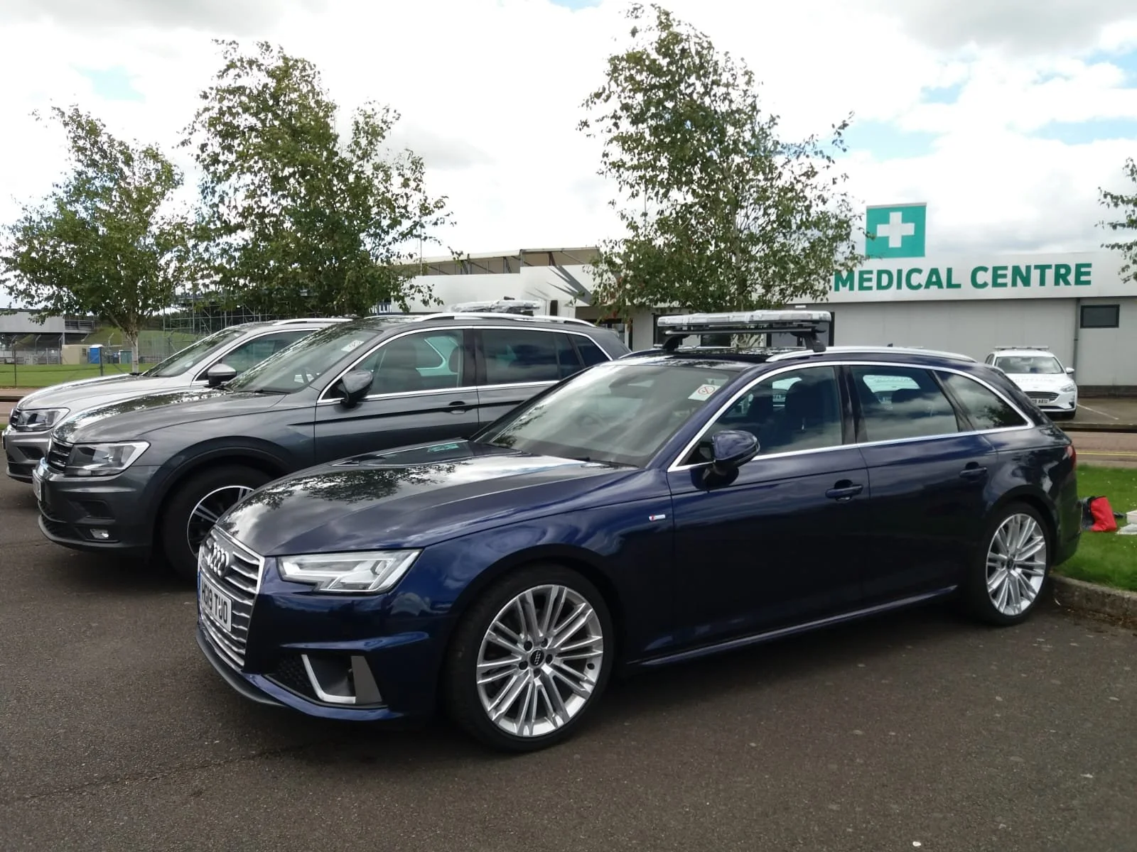 Emergency response vehicles with light bars, including a blue Audi, parked in front of a 'Medical Centre' building at a motorsport venue. There are trees and a cloudy sky in the background.