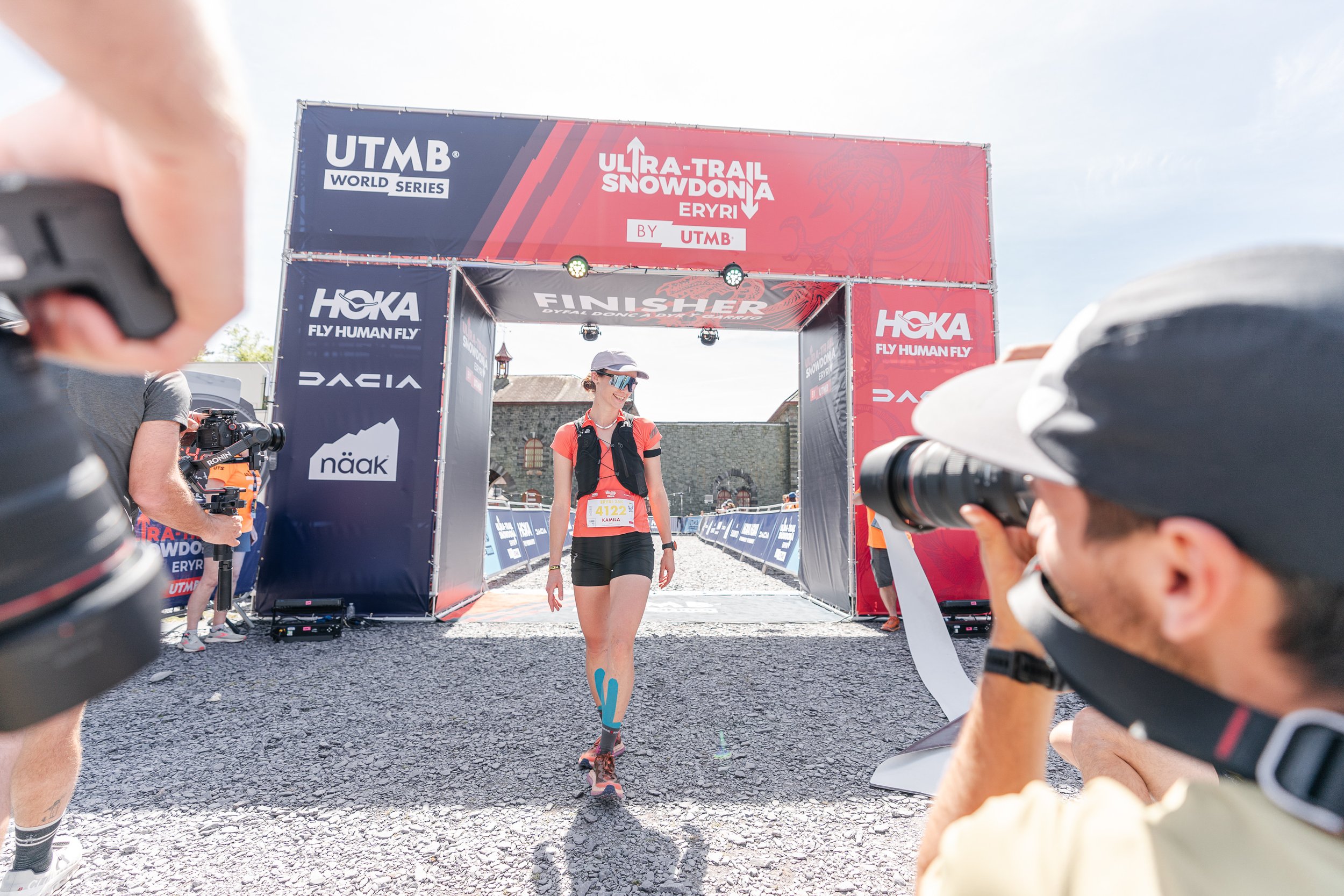 Female athlete crossing the finish line of an Ultra-Trail Snowdonia by UTMB event, smiling, with photographers on either side capturing the moment, and race banners and a structure in the background.