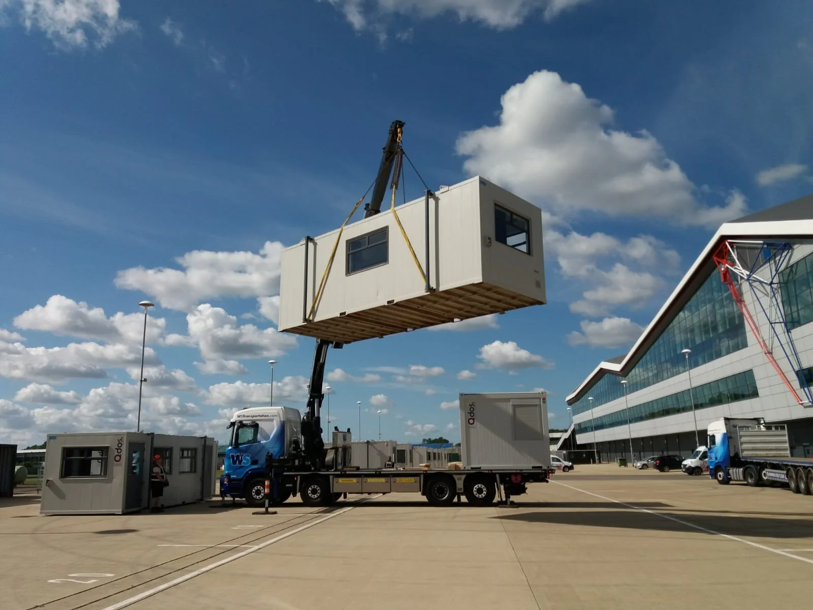A large crane truck is lifting a portable office container high in the air outside a large motorsport building with glass windows. The container is being installed at an outdoor racing paddock with a clear sky and some clouds.