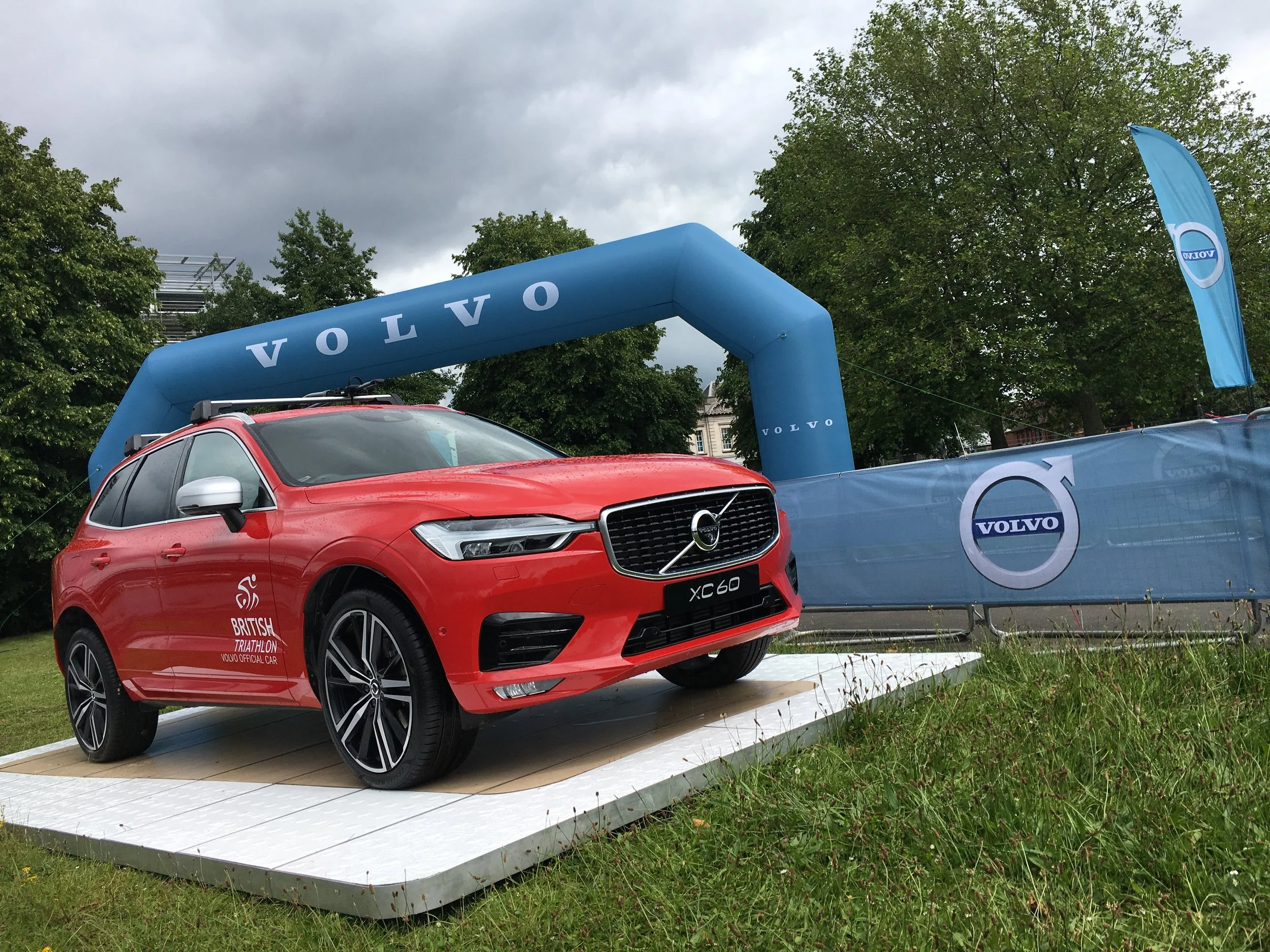 Red Volvo XC60 on display at an outdoor event, with Volvo and British Triathlon logos, under a blue arch branded with 'Volvo' and green trees in the background.
