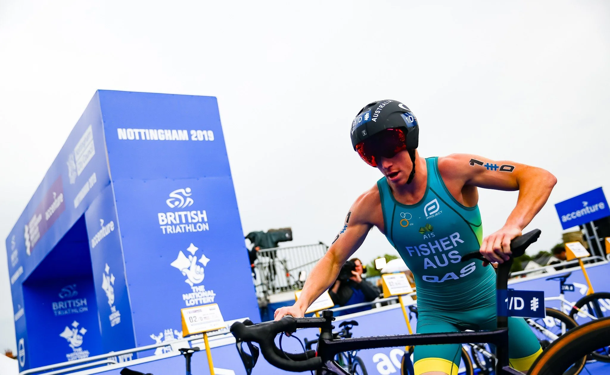 Triathlete in teal and yellow uniform with helmet and sunglasses, pushing a bicycle in transition at the Nottingham 2019 World Triathlon event.