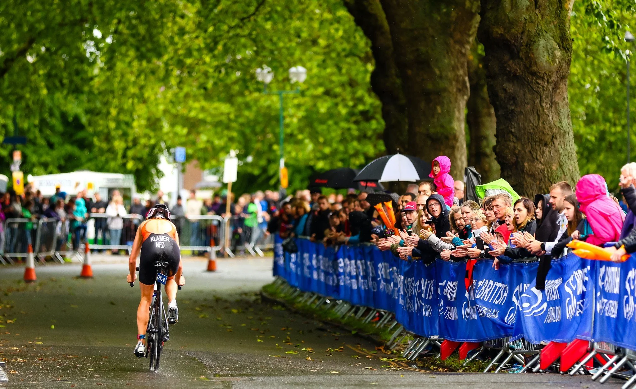 A female athlete bikes along a wet, tree-lined street during a race while crowd spectators cheer from behind blue barriers in the rain, some holding umbrellas and children in raincoats.