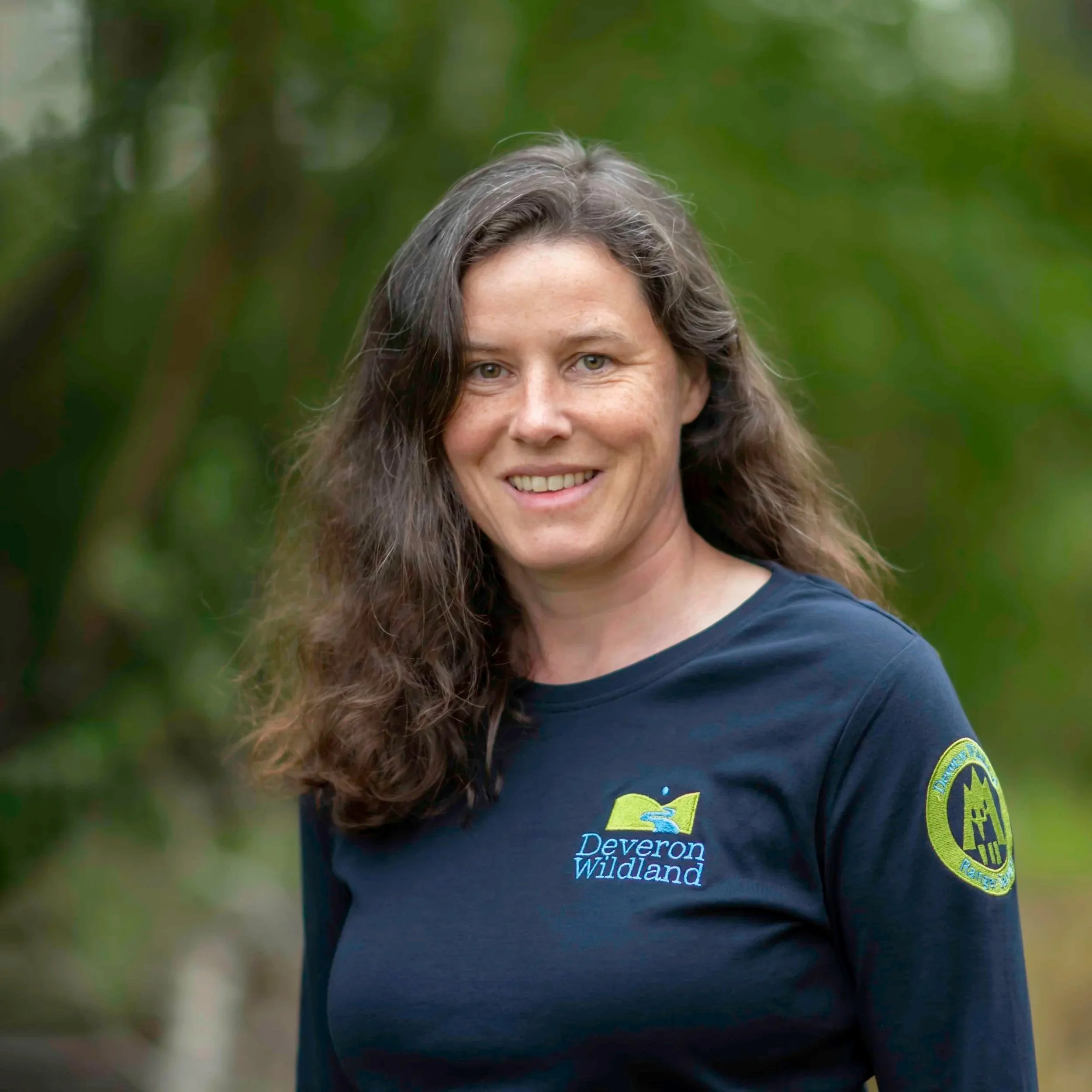 A woman with long wavy brown-grey hair and light skin, smiling outdoors with a blurred green background. She is wearing a blue T-shirt with text "Deveron Wildlands" and a patch on the sleeve.