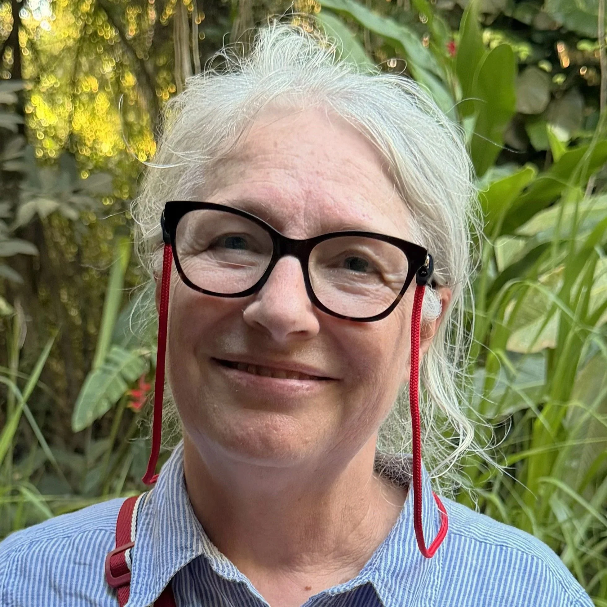 Close-up of a smiling  woman with white hair and glasses, surrounded by green foliage, wearing a blue striped shirt.