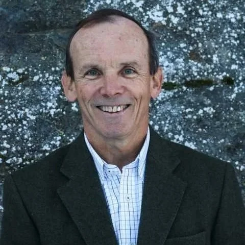 Smiling man in a suit jacket and collared shirt standing outdoors with a wall in the background.