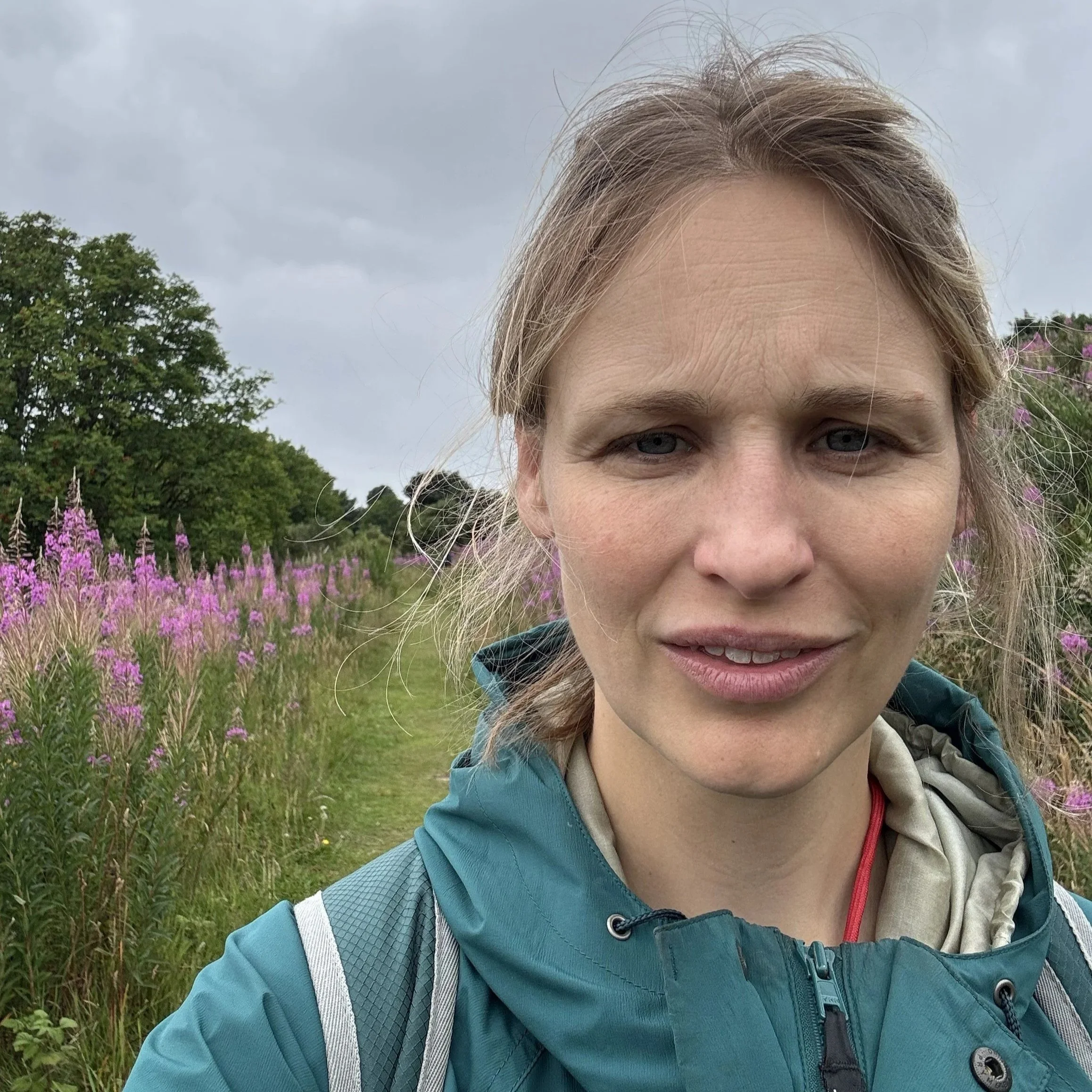 Close-up of a woman with blonde hair and blue eyes outdoors, wearing a teal and grey jacket, with pink flowers and green bushes in the background under a cloudy sky.