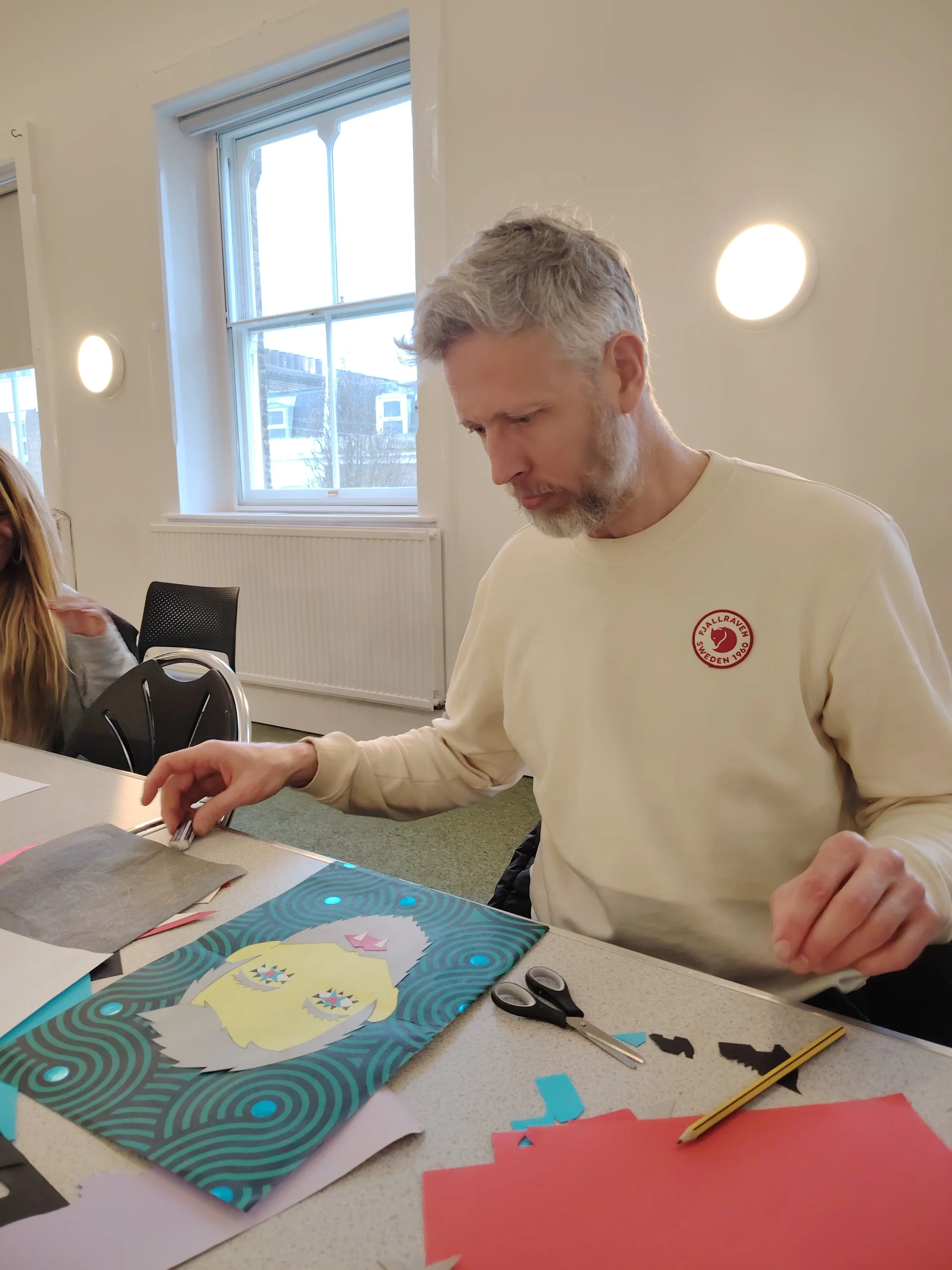A man with gray hair and beard wearing a cream-colored sweatshirt with a red circular logo, sitting at a table working on a paper craft project. The table has scissors, paper, and cut-out shapes, with a colorful paper featuring a face with yellow hair, pink bow, and starry eyes. The background shows a room with white walls, a window, and modern lighting.