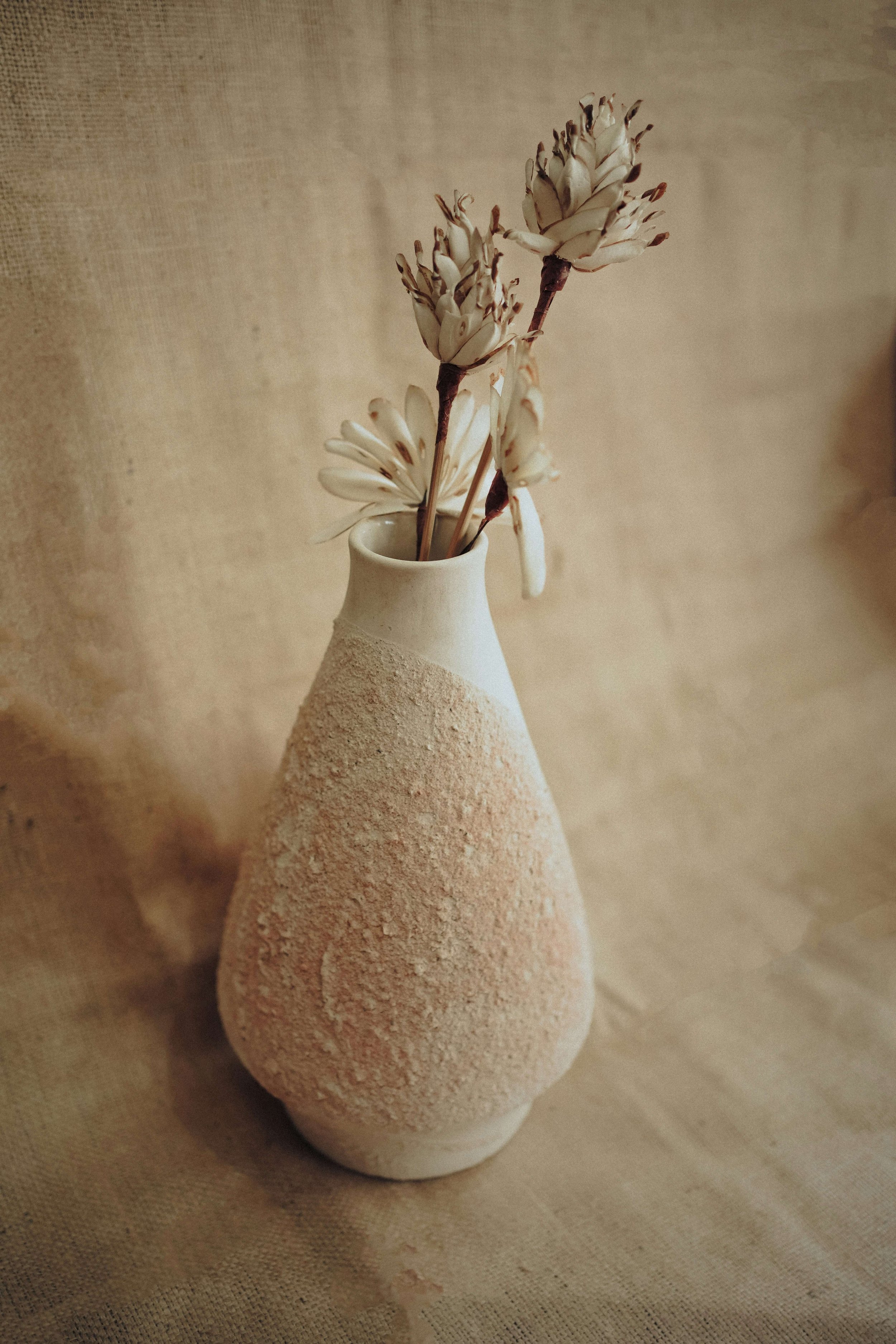 Ceramic vase with textured surface holding dried flowers, set against beige fabric background.