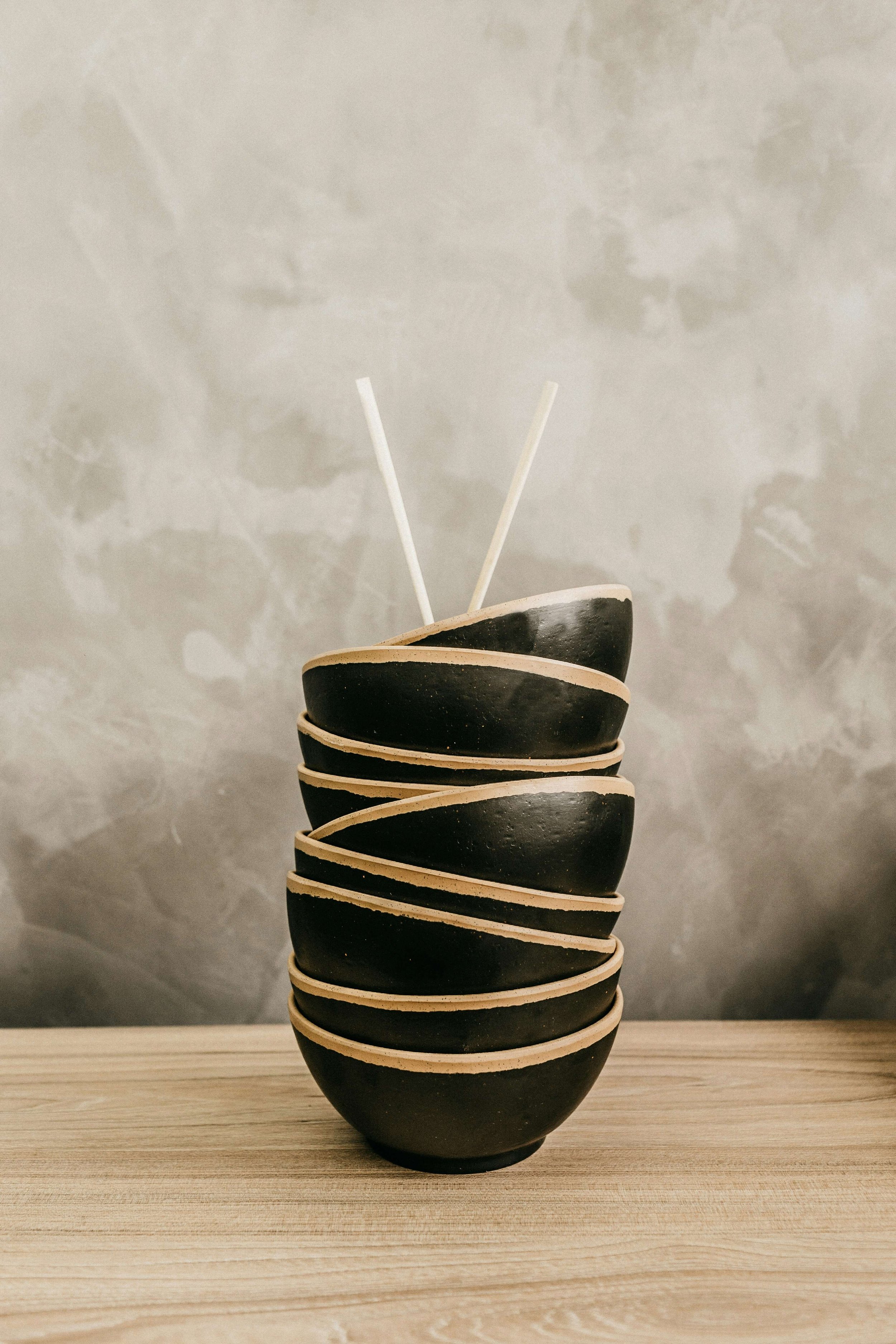 Stack of black bowls with cream-colored edges and two white sticks placed inside, on a wooden surface against a textured gray wall.