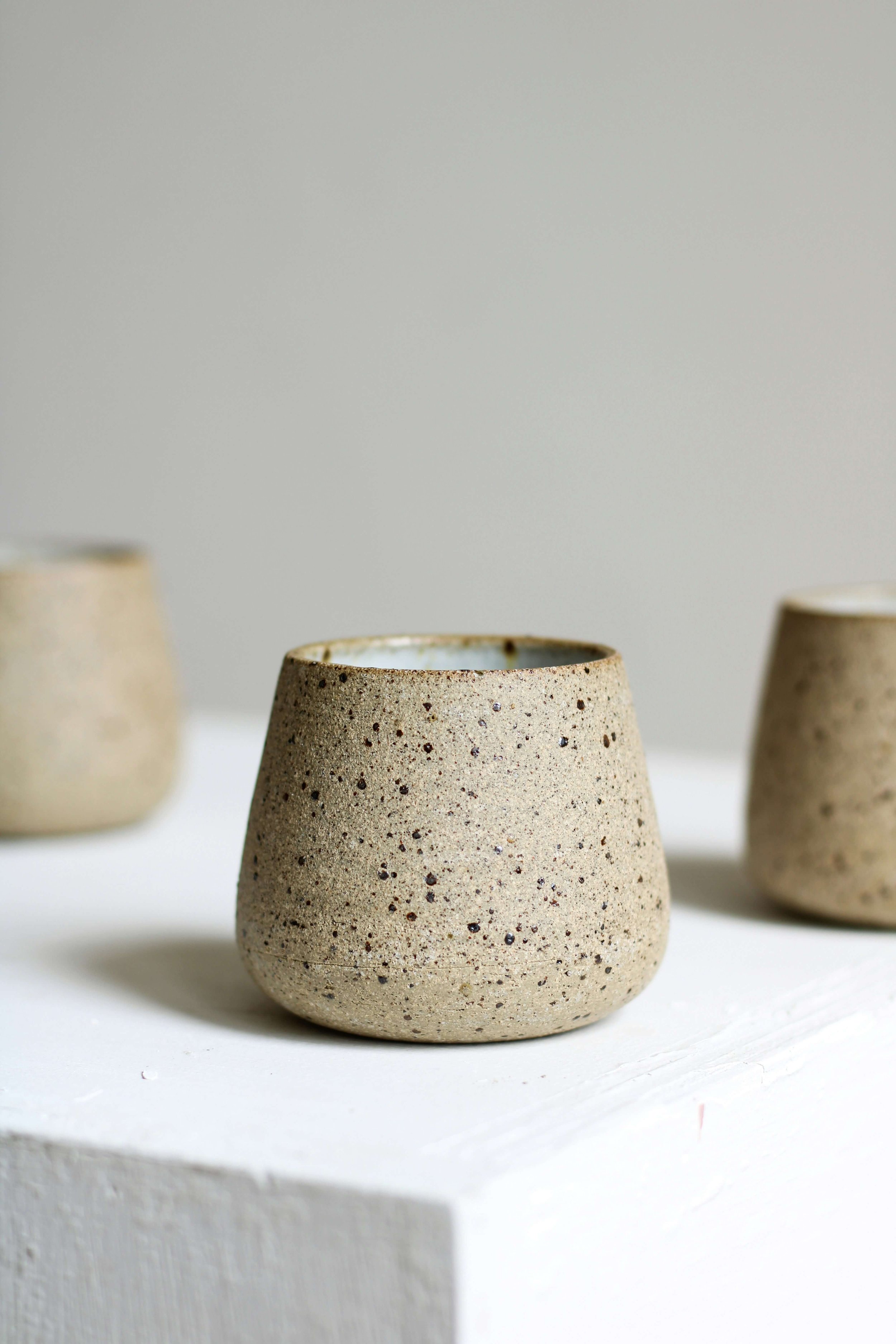 Three beige speckled ceramic cups on a white surface against a plain grey background.