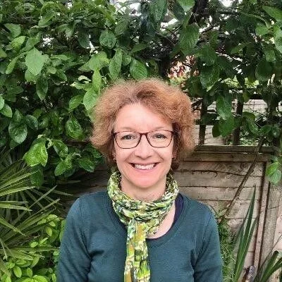 A woman with glasses and curly hair smiling outdoors, standing in front of lush green foliage and a wooden fence.