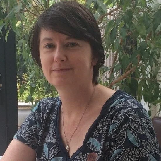 A woman with short dark hair, wearing a dark blue top with a leaf pattern, sitting outdoors near green foliage.