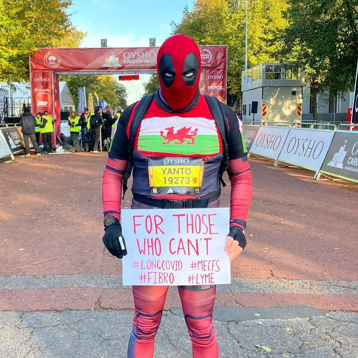 A person dressed in Deadpool costume holding a sign at a marathon event, with others and event banners in the background.