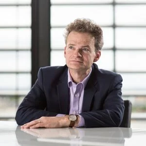 A middle-aged man in a dark suit and light purple shirt sitting at a white table indoors, looking thoughtful.