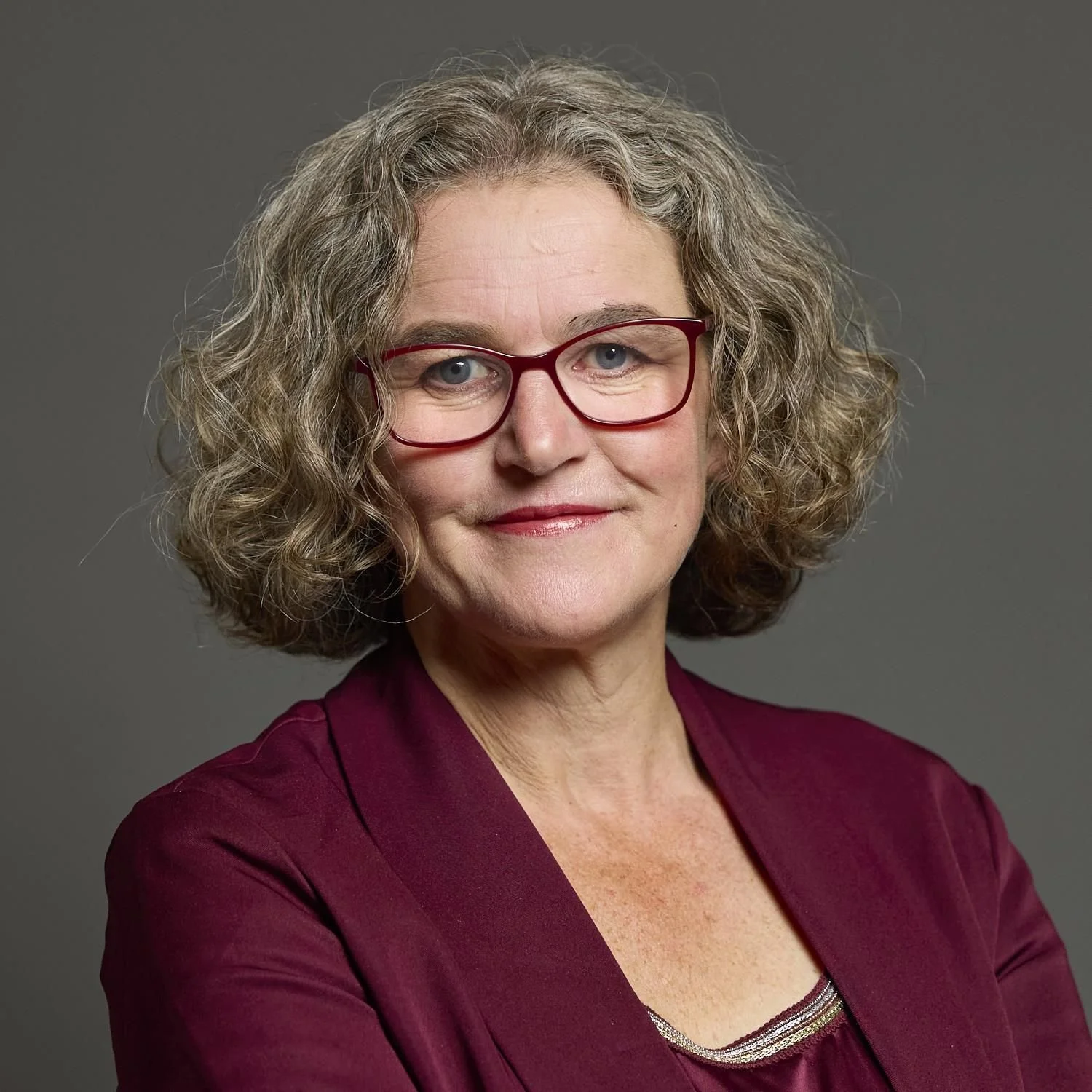 A portrait of a middle-aged woman with curly gray hair, wearing red glasses, a maroon blouse, and a subtle smile against a gray background.
