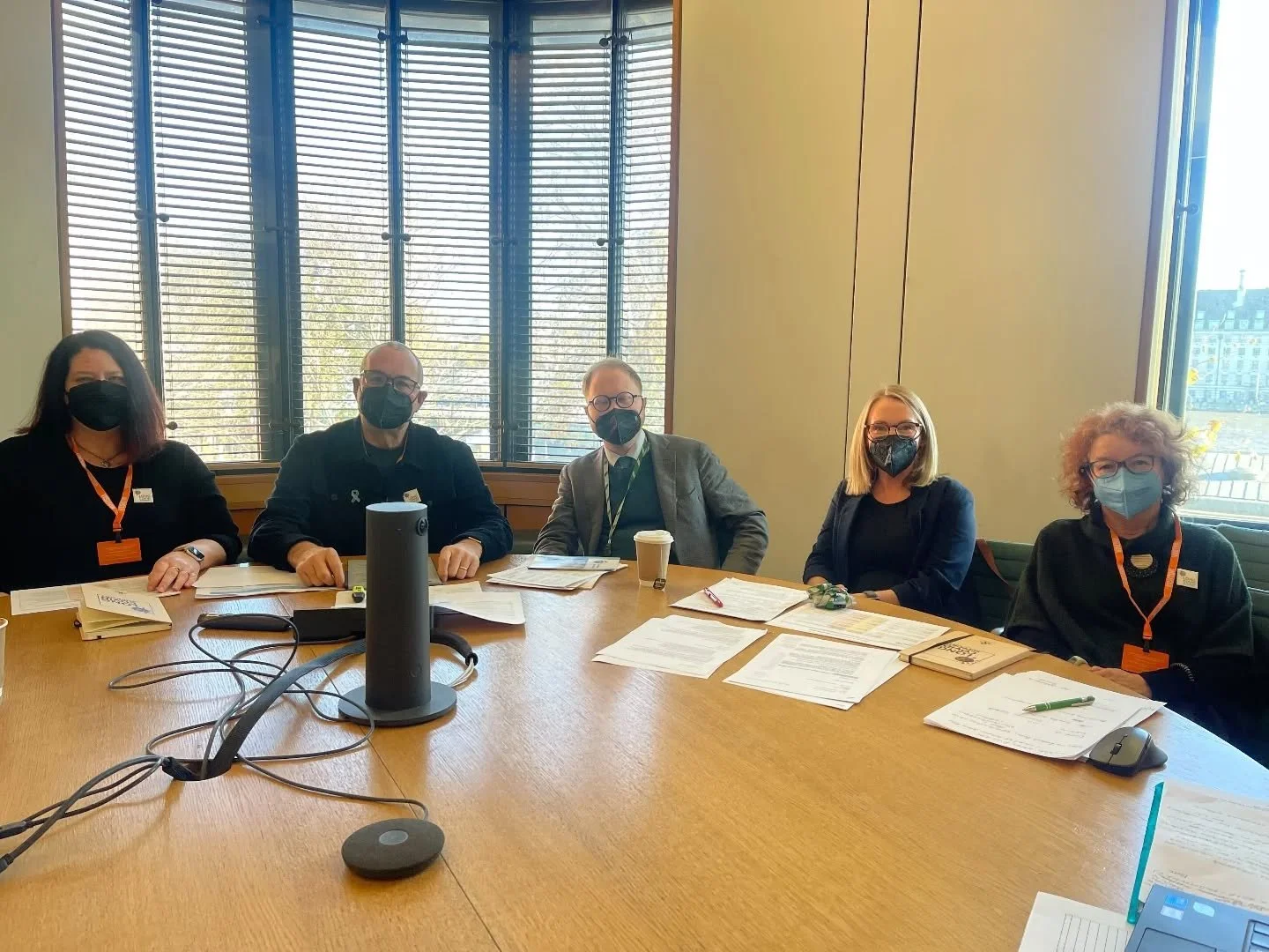 Five individuals sitting at a conference table wearing masks, with documents, books, a coffee cup, and electronic devices on the table, in a room with large windows and blinds.