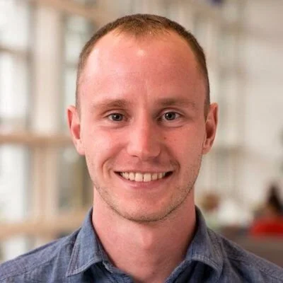 Portrait of a young man with short hair, smiling, wearing a blue collared shirt, indoors with a blurred background.