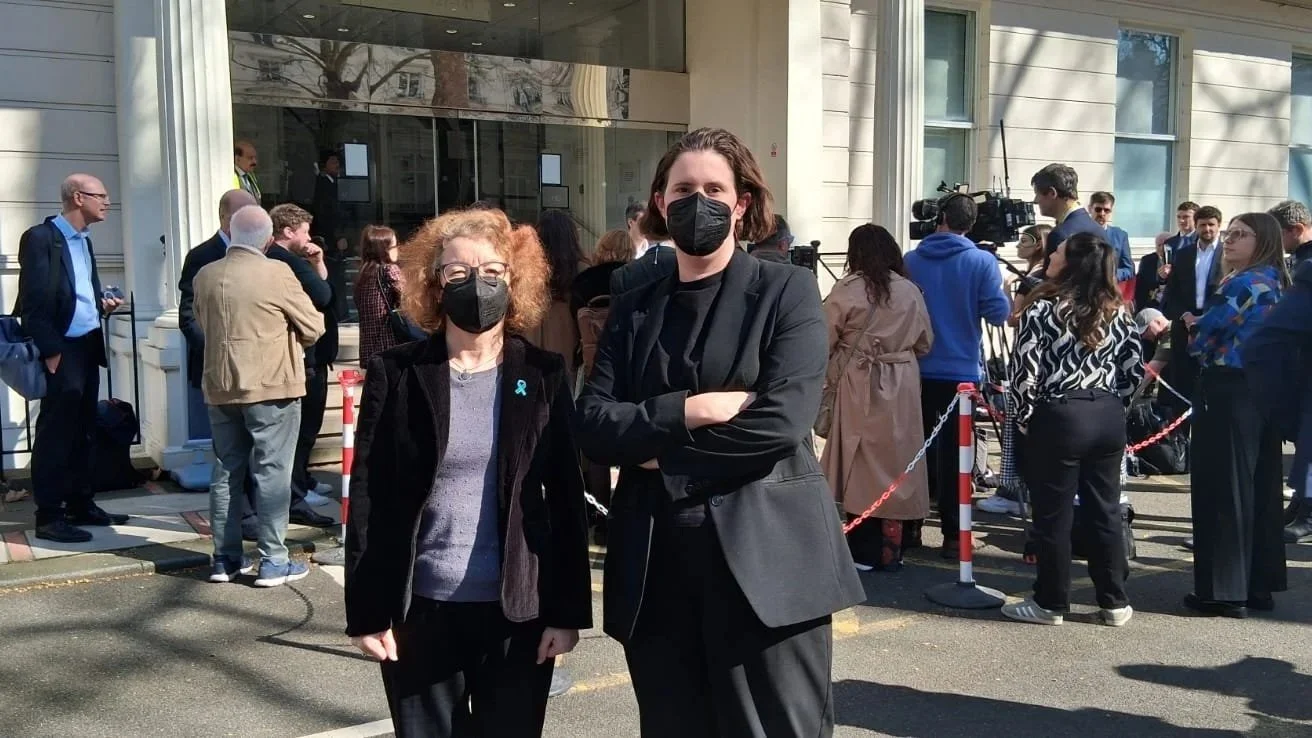 Two women wearing face masks standing outside a building with a crowd in the background, some filming with cameras.