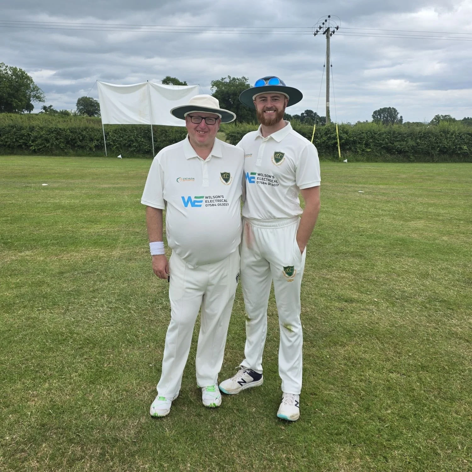 Two men dressed in white cricket uniforms with hats, standing on a cricket field, smiling for the camera. There are cricket equipment and a wicket keeper's stand in the background, with cloudy skies overhead.
