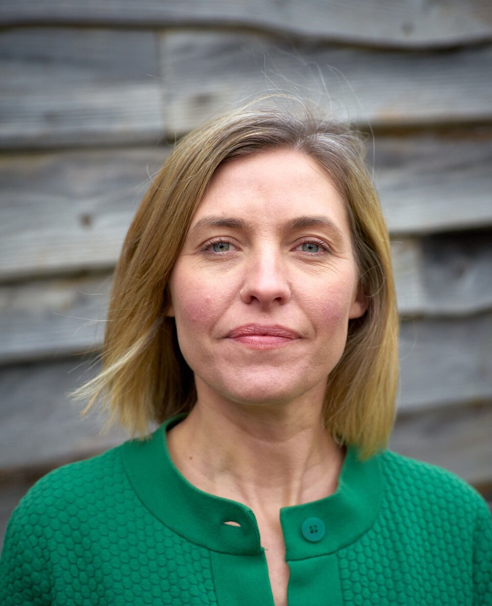 A woman with blonde hair and blue eyes wearing a green textured blazer standing in front of a wooden wall.