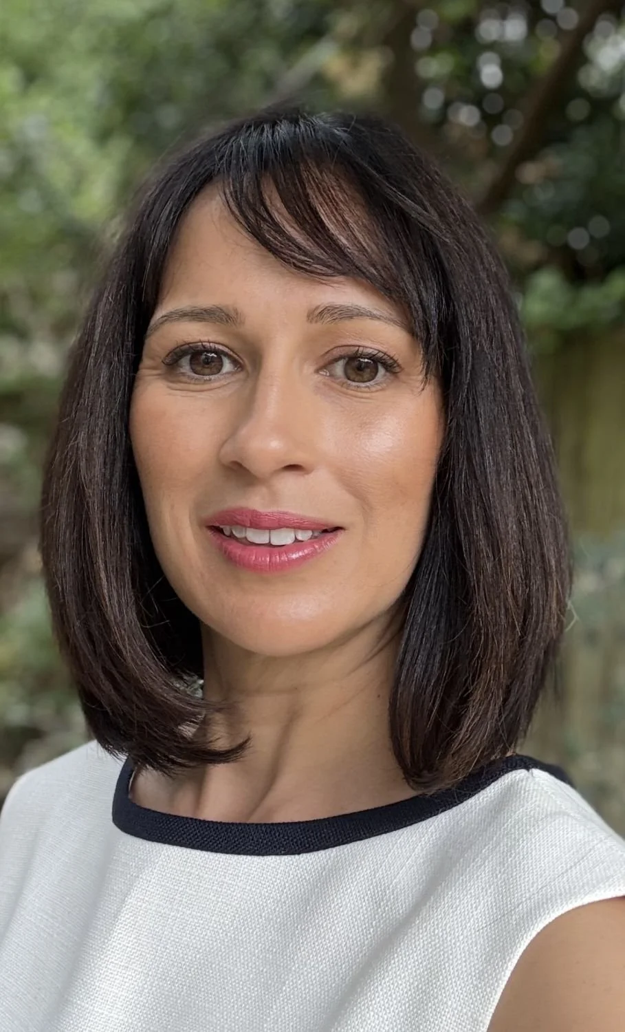 Close-up of a woman with shoulder-length dark brown hair, wearing a white top with black trim, smiling outdoors with blurred green trees in the background.